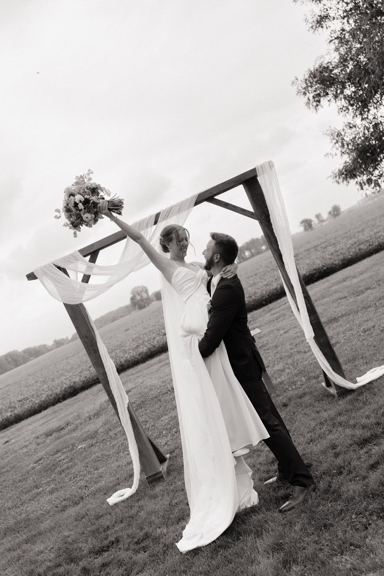 Black and white photo of bride and groom embracing under rustic wooden arch with flowing ribbons in open field.