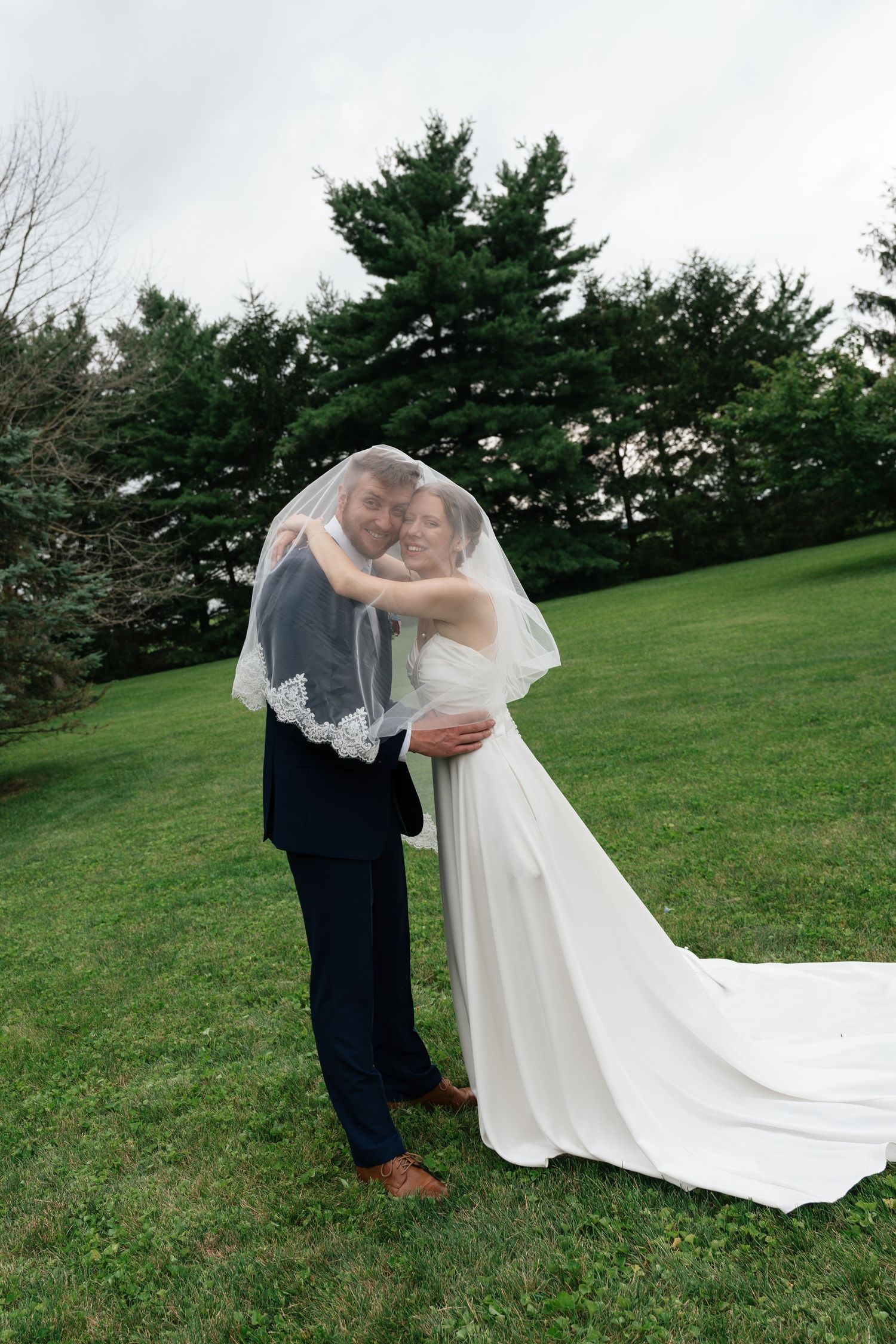 Bride and groom share a romantic moment under wedding veil on green lawn with evergreen trees backdrop.