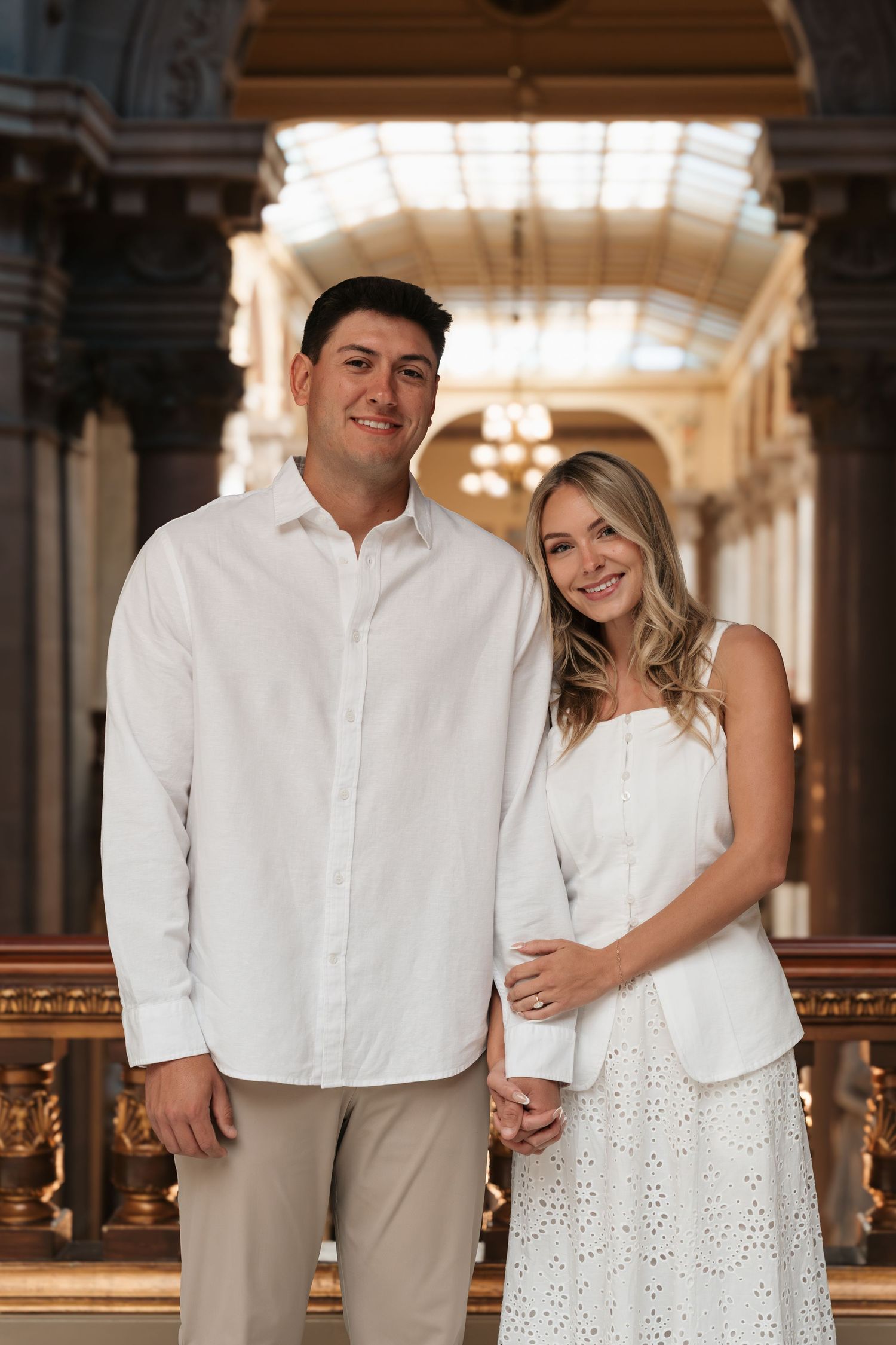 Couple in matching white outfits posing together in an elegant ornate hall with chandeliers.