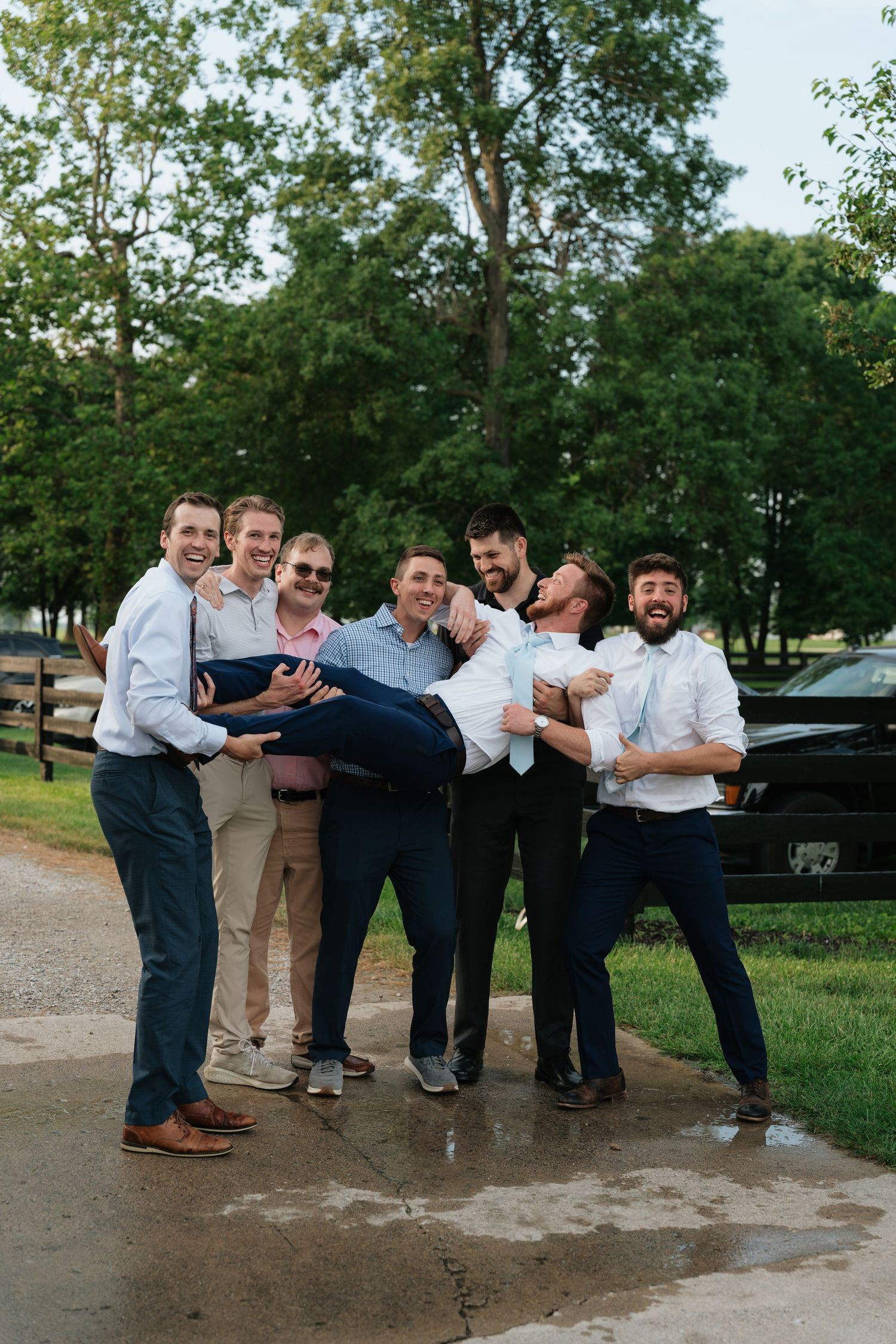 Groomsmen laughing and carrying groom outdoors at wedding celebration near wooden fence and green trees.