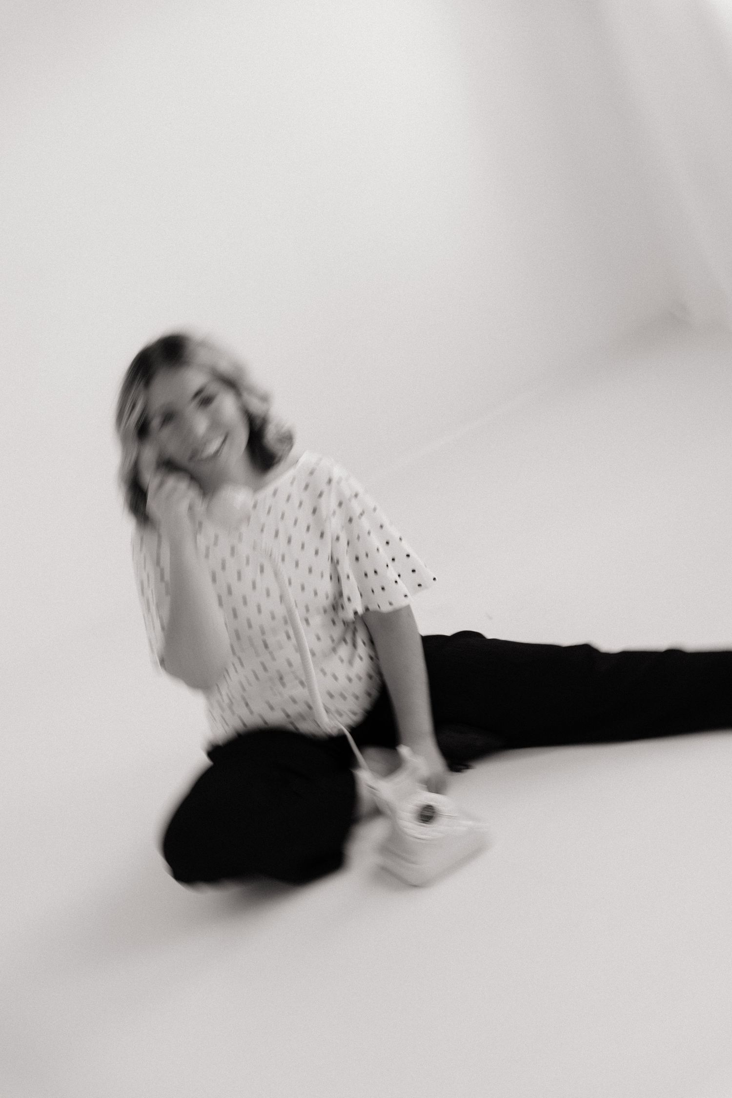 Black and white photo of a smiling young woman sitting on a white studio floor in a patterned top and dark pants.
