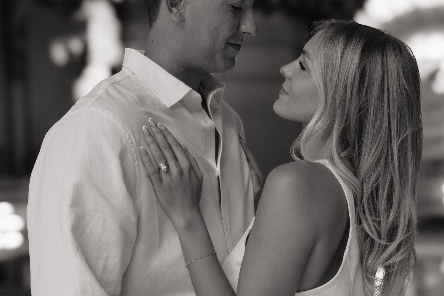 Black and white photo of a couple embracing romantically, woman wearing engagement ring, smiling at each other.