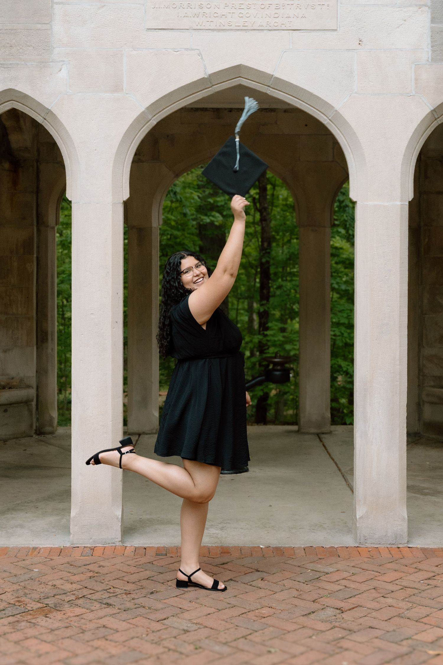 A joyful graduate in a black dress raises her cap triumphantly under stone arches on a brick courtyard.