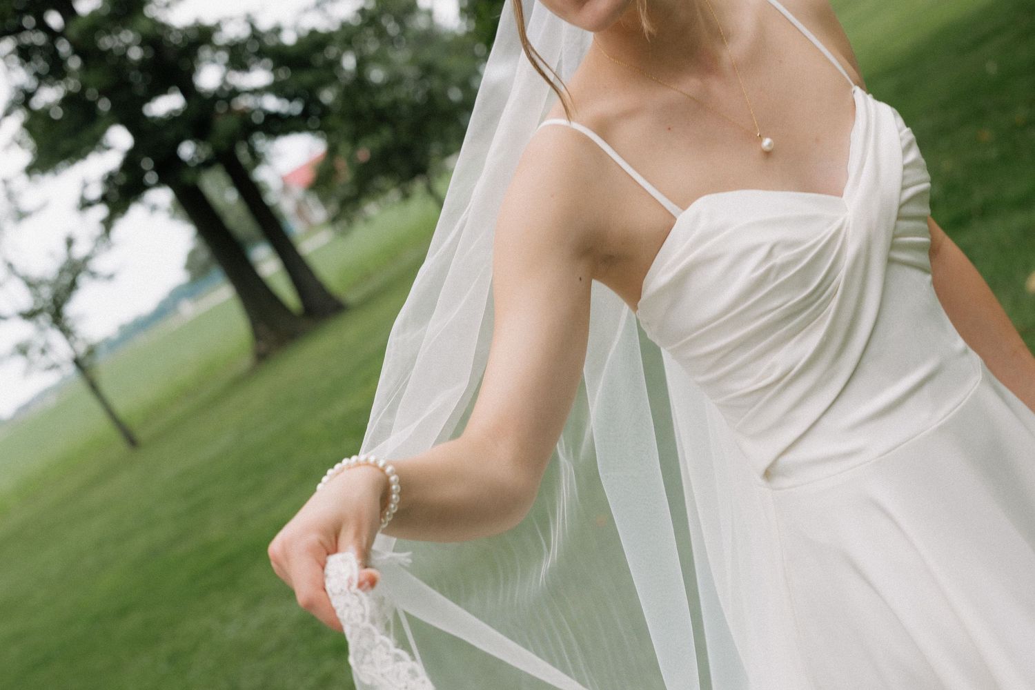 Bride in white gown holding lace veil, wearing pearl bracelet and necklace, standing on green lawn.