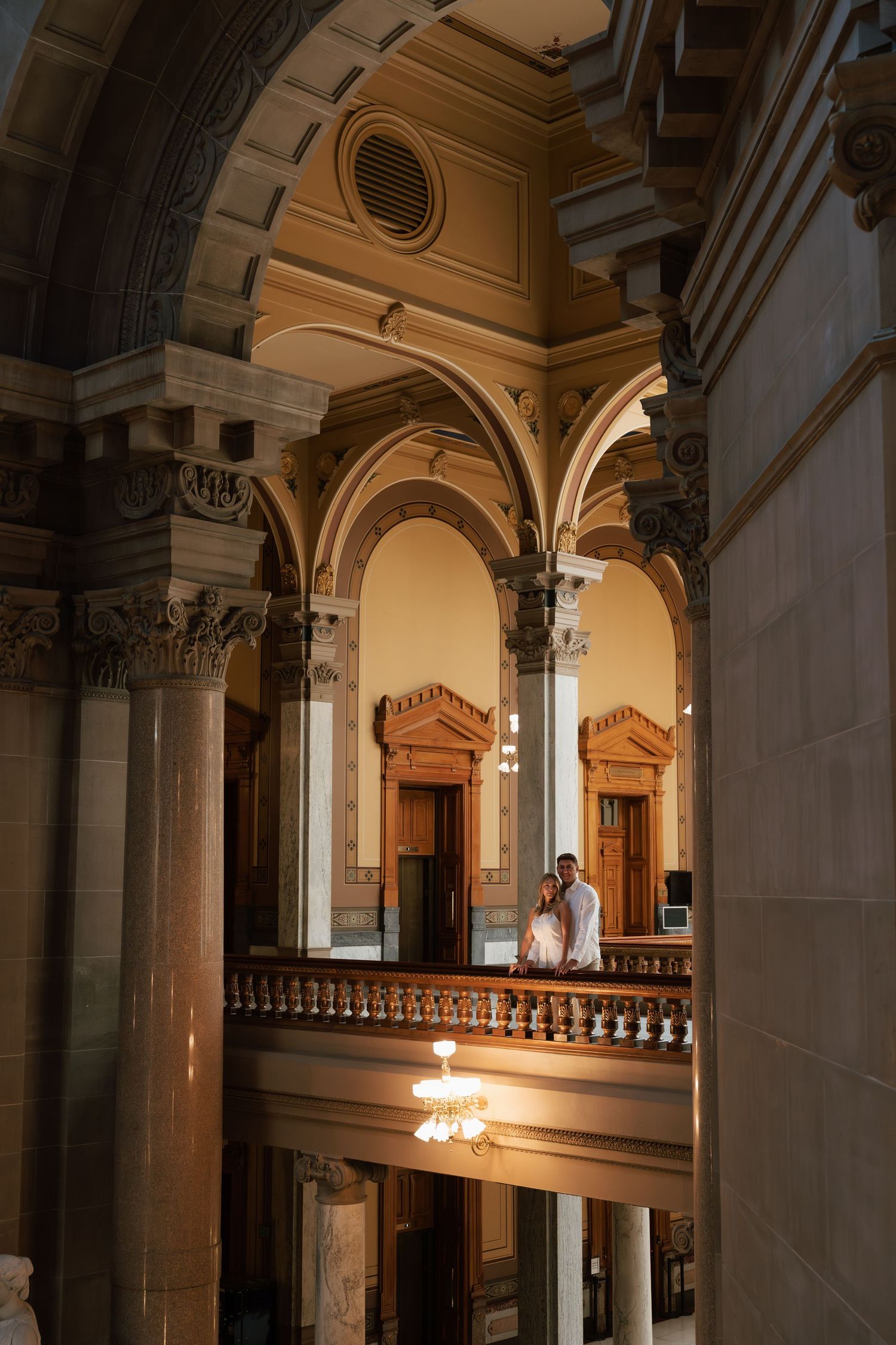 Couple embracing on ornate balcony inside grand historic building with arched ceilings and classical columns.