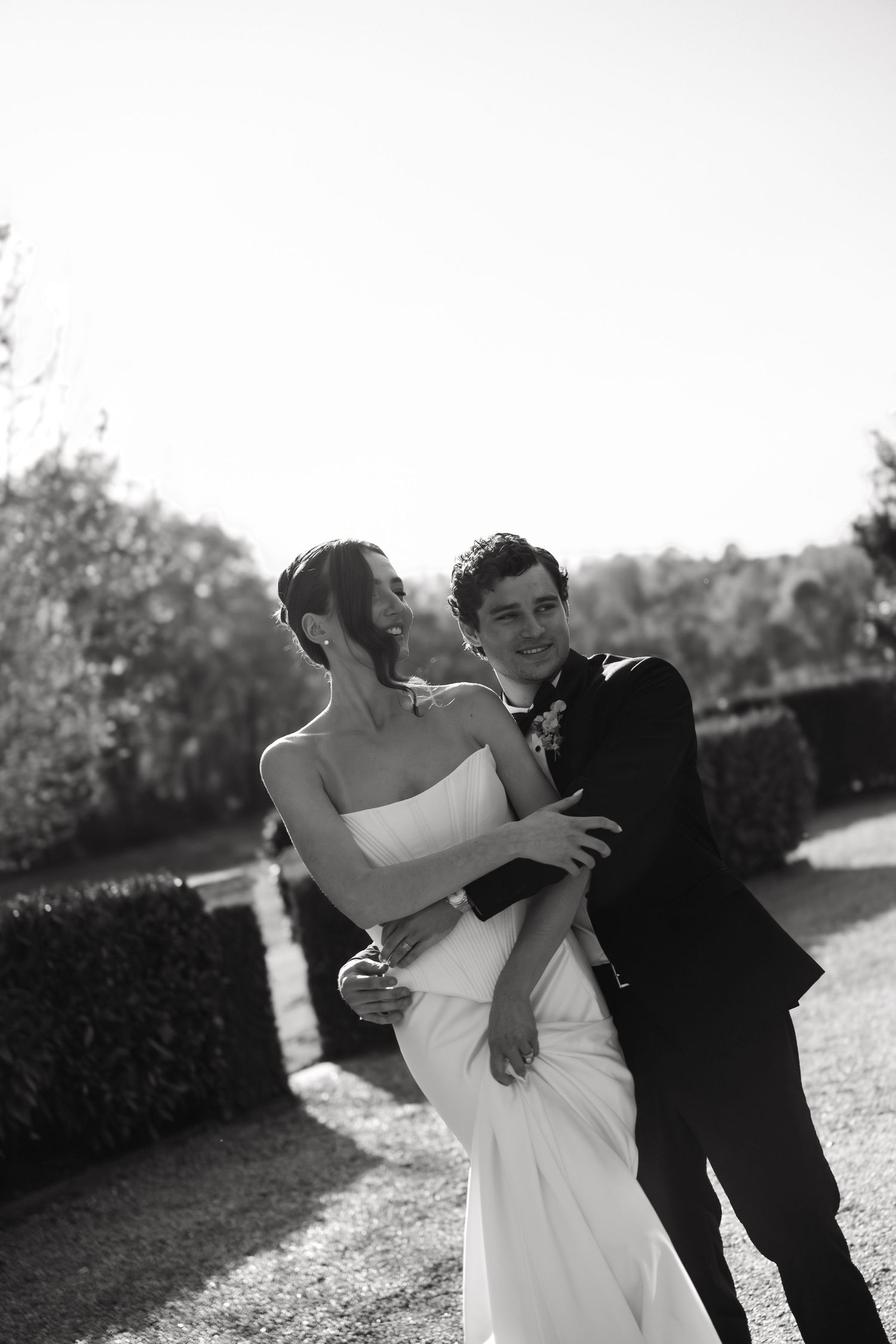 Black and white photo of a bride and groom embracing and smiling outdoors at a garden wedding venue.