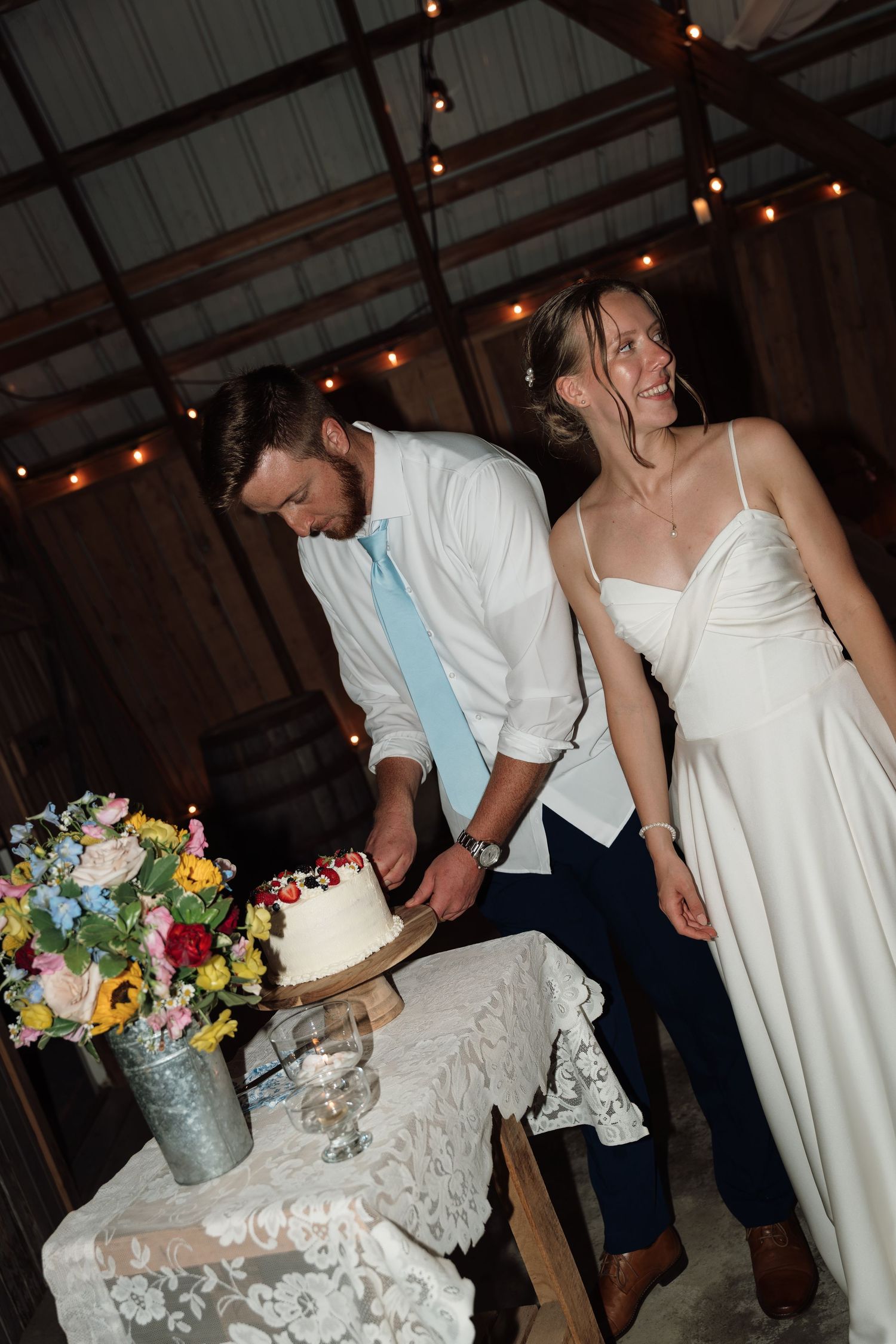 Bride and groom cutting white wedding cake in rustic barn with string lights.