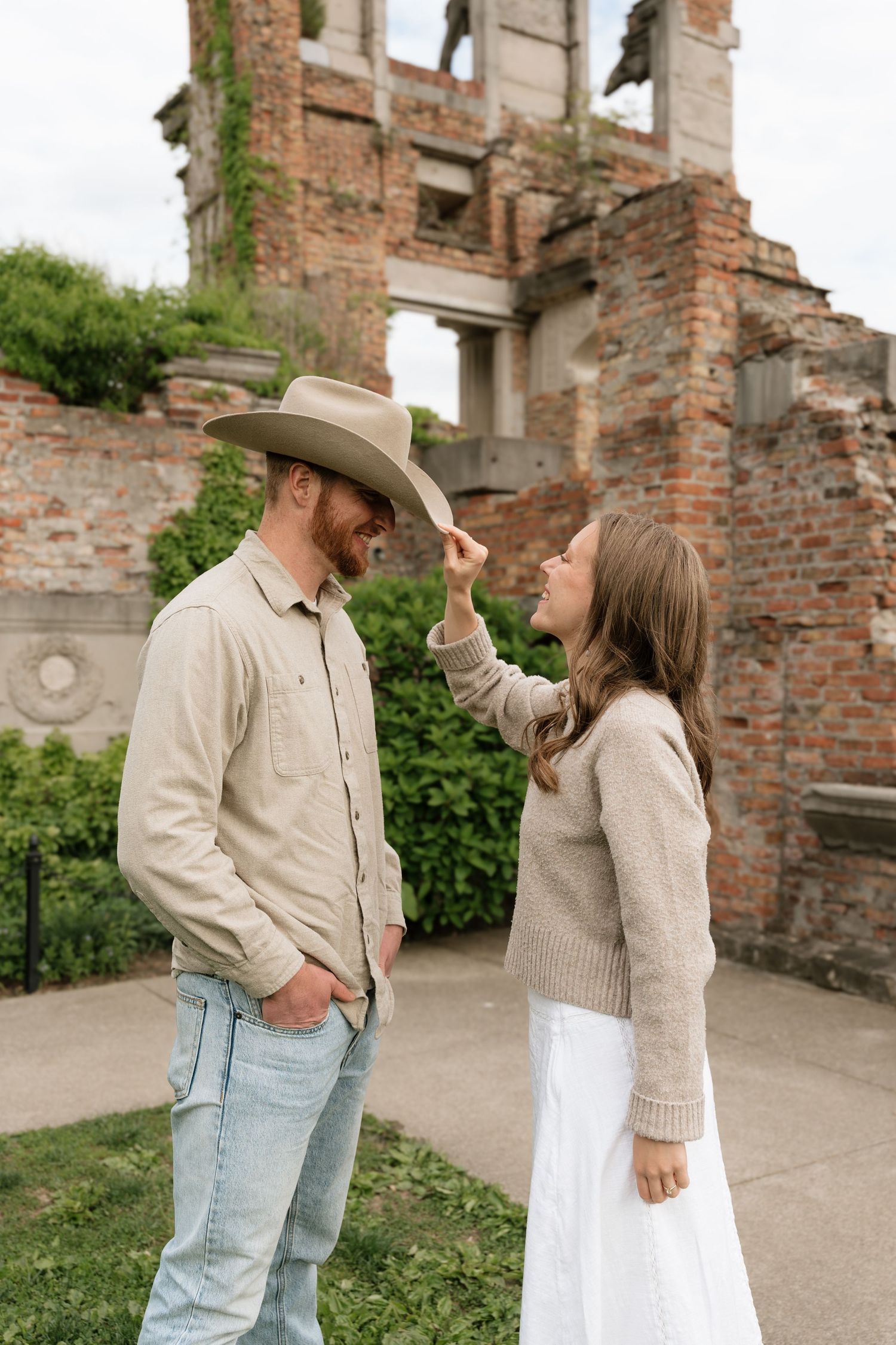 Woman playfully tipping mans cowboy hat near ivy-covered brick ruins in a garden.