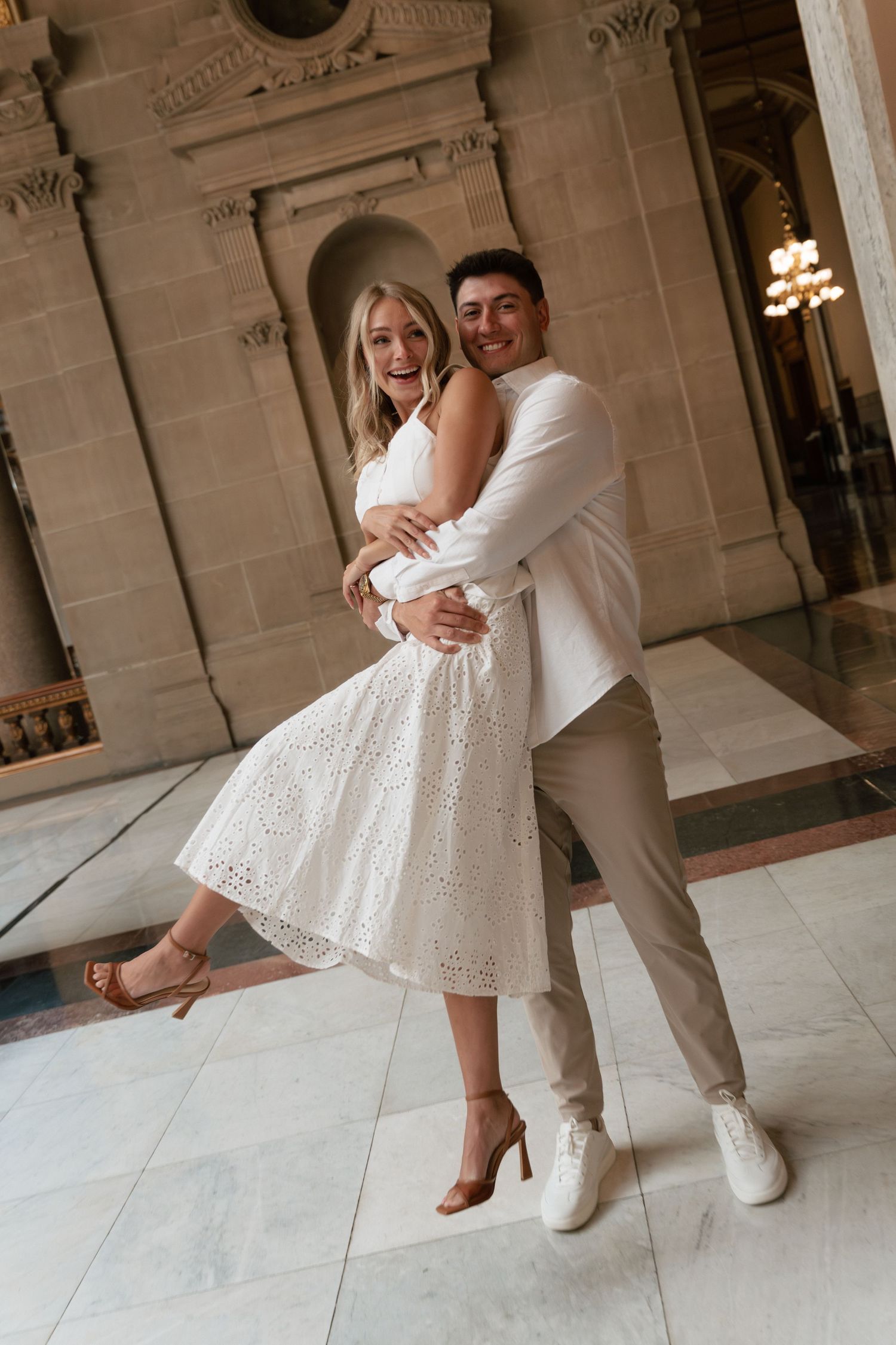 Joyful couple embracing and laughing in elegant marble hall, woman in white eyelet dress, man in white blazer.