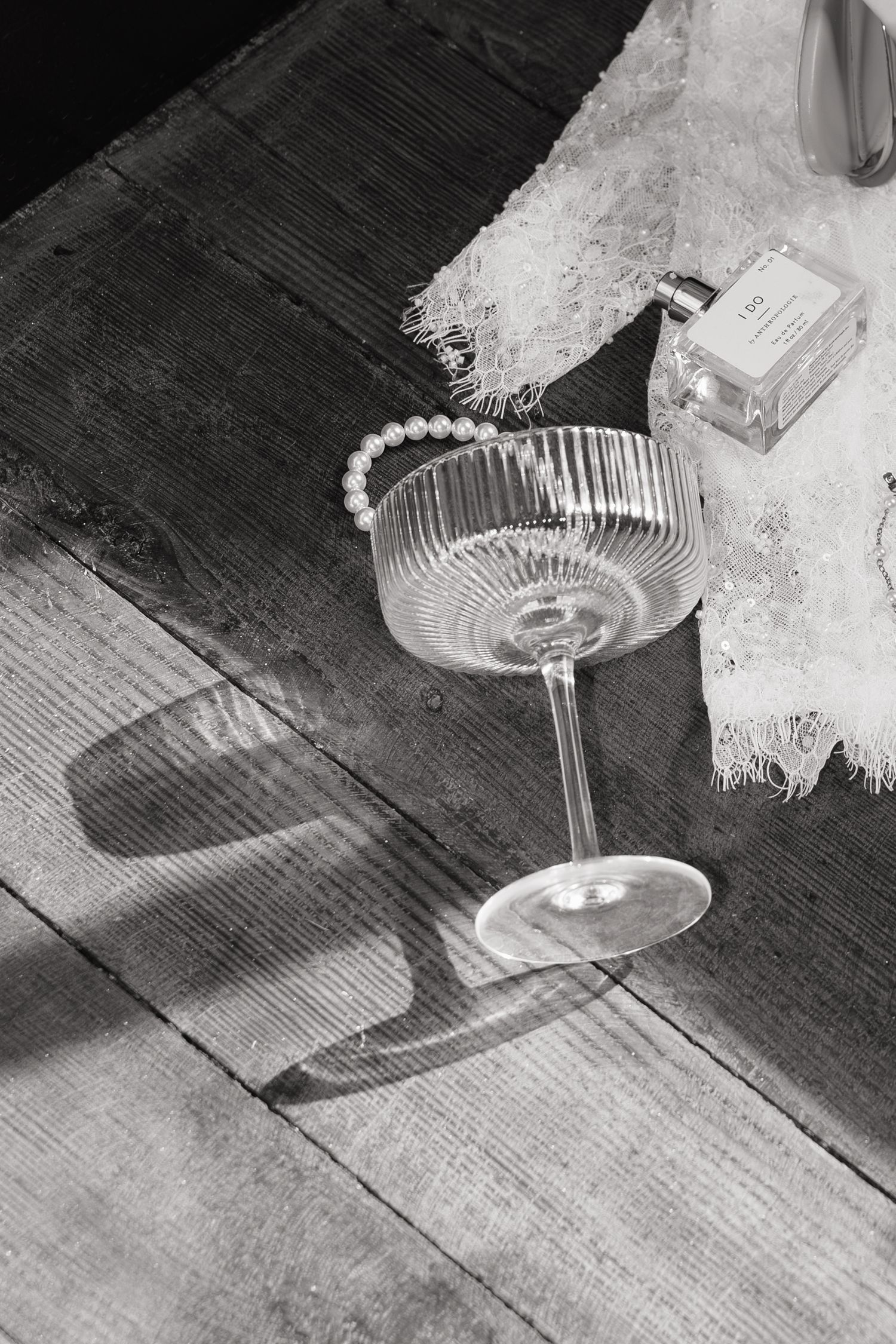Black and white photo of ribbed coupe glass with pearl bracelet on wooden surface beside perfume bottle.