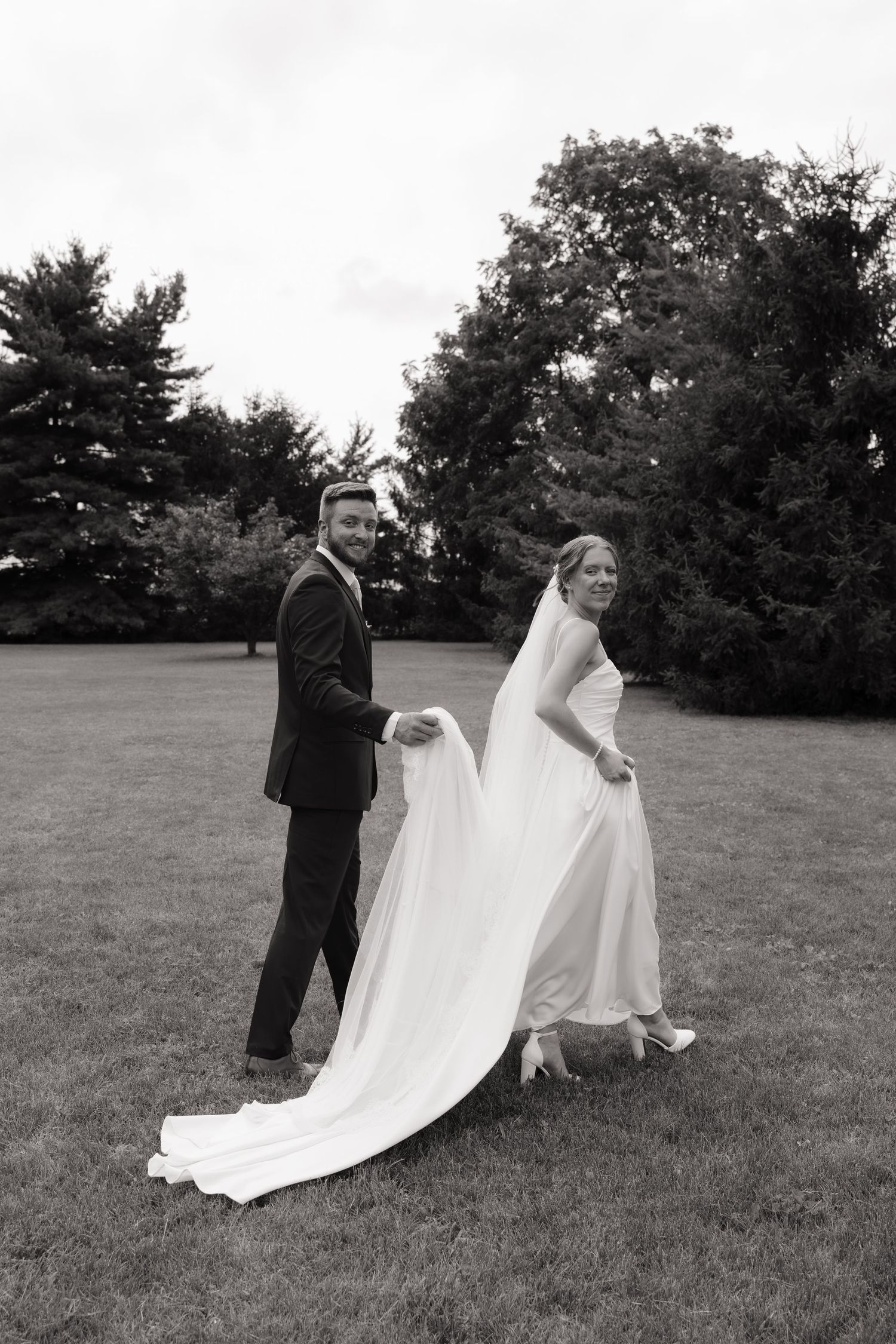 Black and white photo of a groom holding his bride's wedding dress train as they walk across a green lawn.