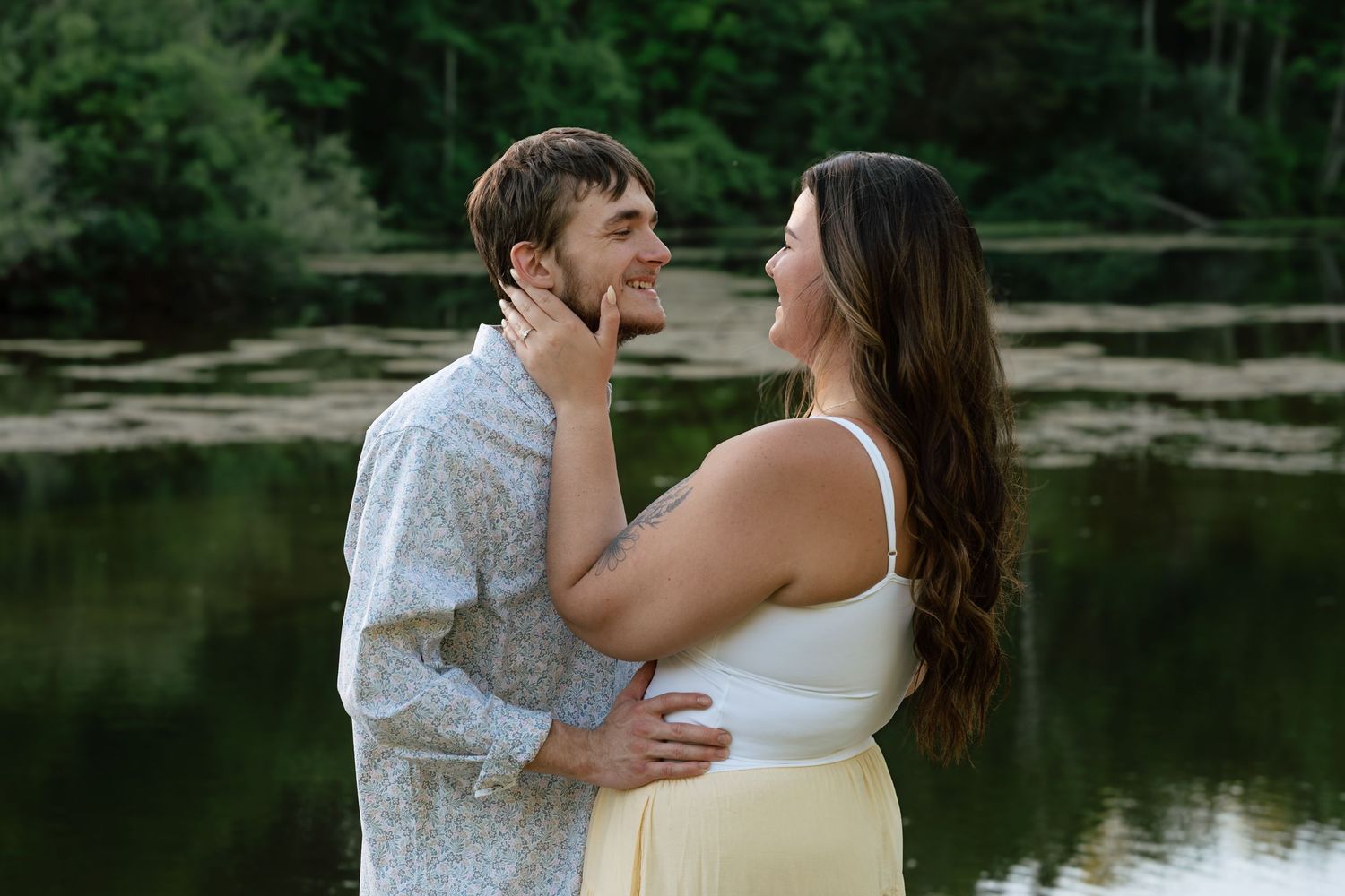 Couple sharing a romantic moment by a serene forest lake, woman in white top and yellow skirt touching mans face.