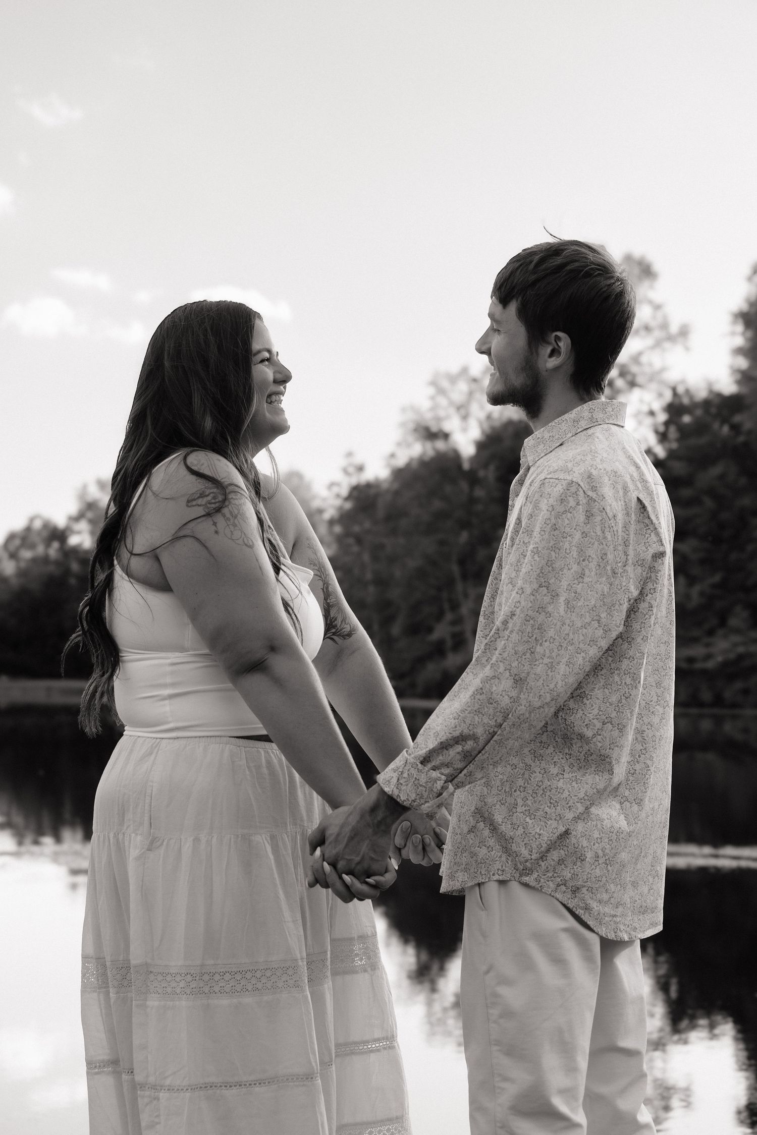 Black and white photo of a couple holding hands and smiling at each other near a reflective lake with trees in background.