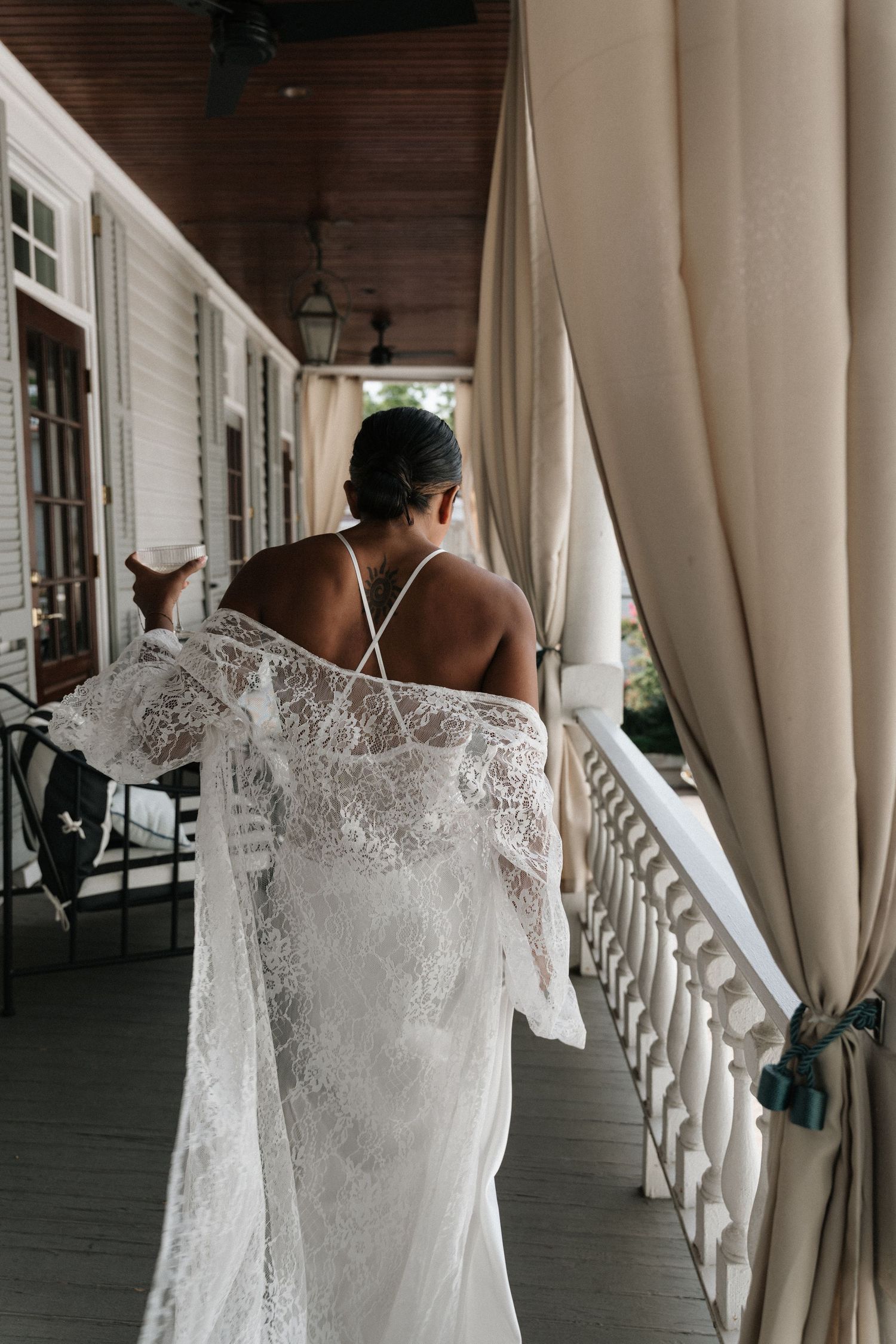 Bride in elegant lace wedding gown stands on a classic Southern porch, viewed from behind, holding her flowing veil.