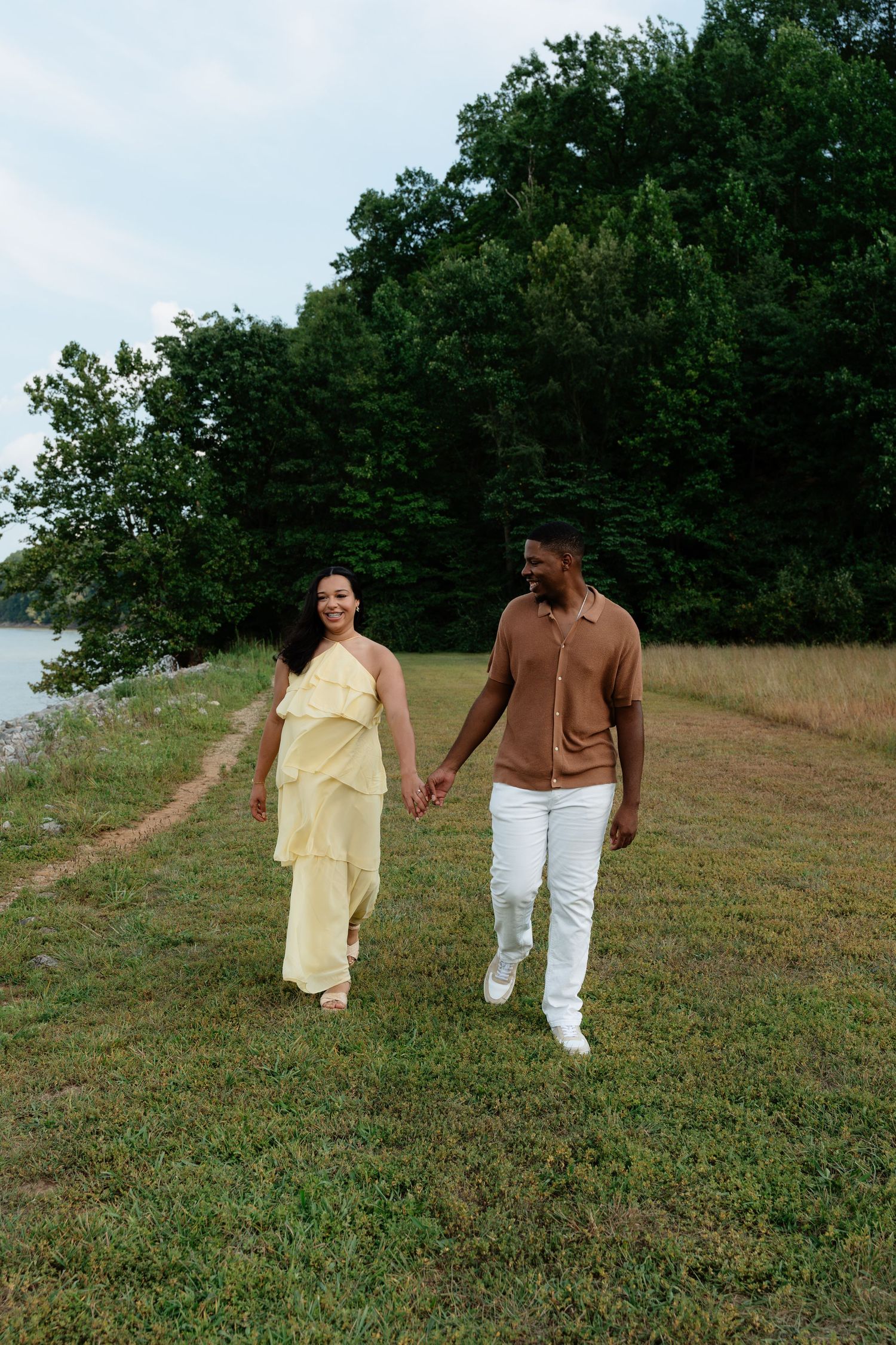 A couple holds hands walking through a green field near a lake, woman in yellow dress, man in brown shirt and white pants.