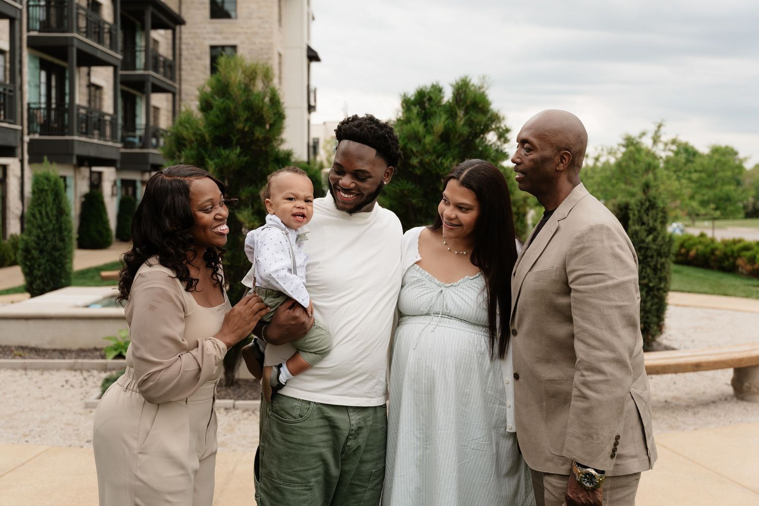 A joyful multigenerational family poses outdoors, with a man holding a baby, beside a pregnant woman and smiling relatives.