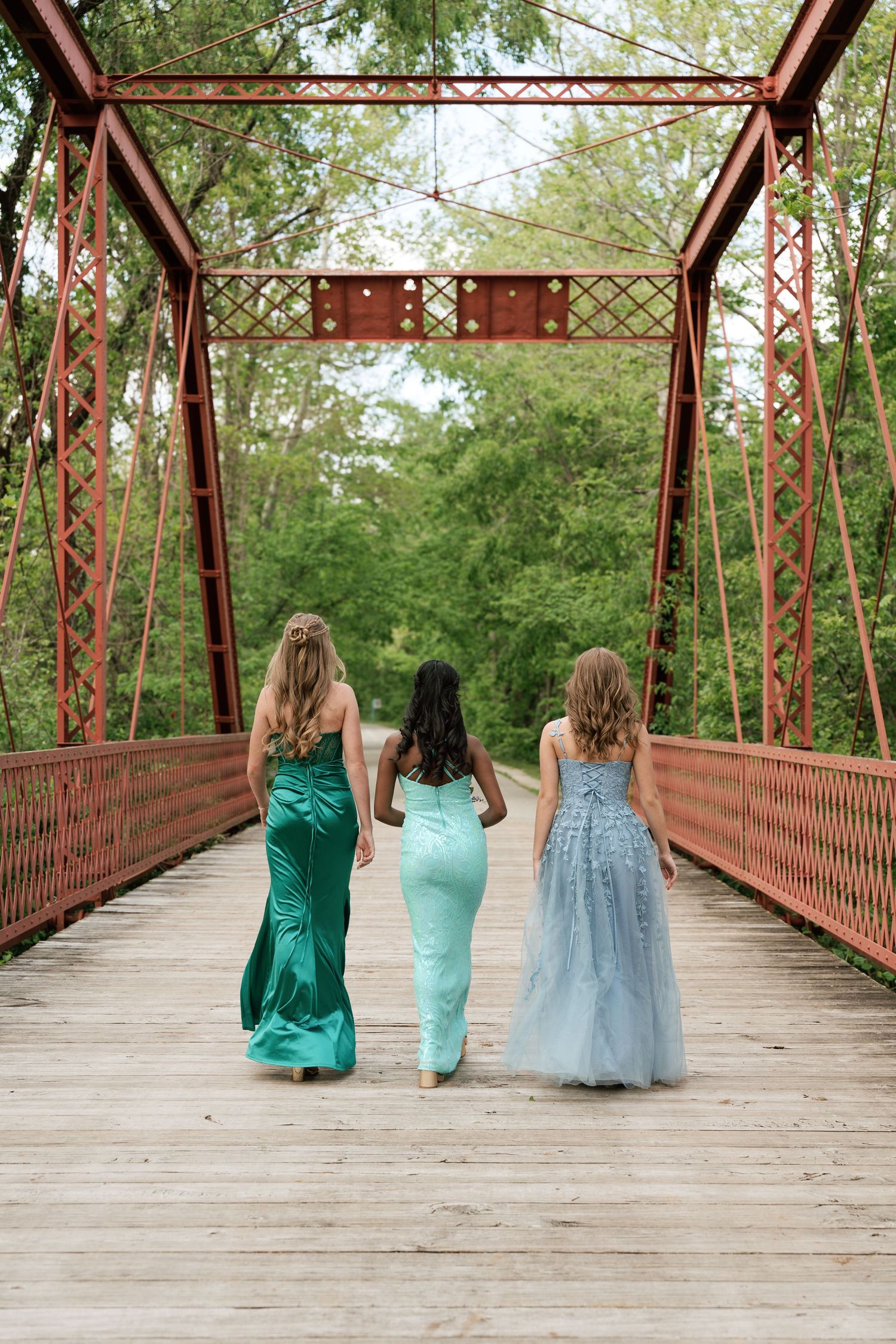 Three girls in elegant prom dresses walk across a rustic red iron bridge surrounded by lush green trees.