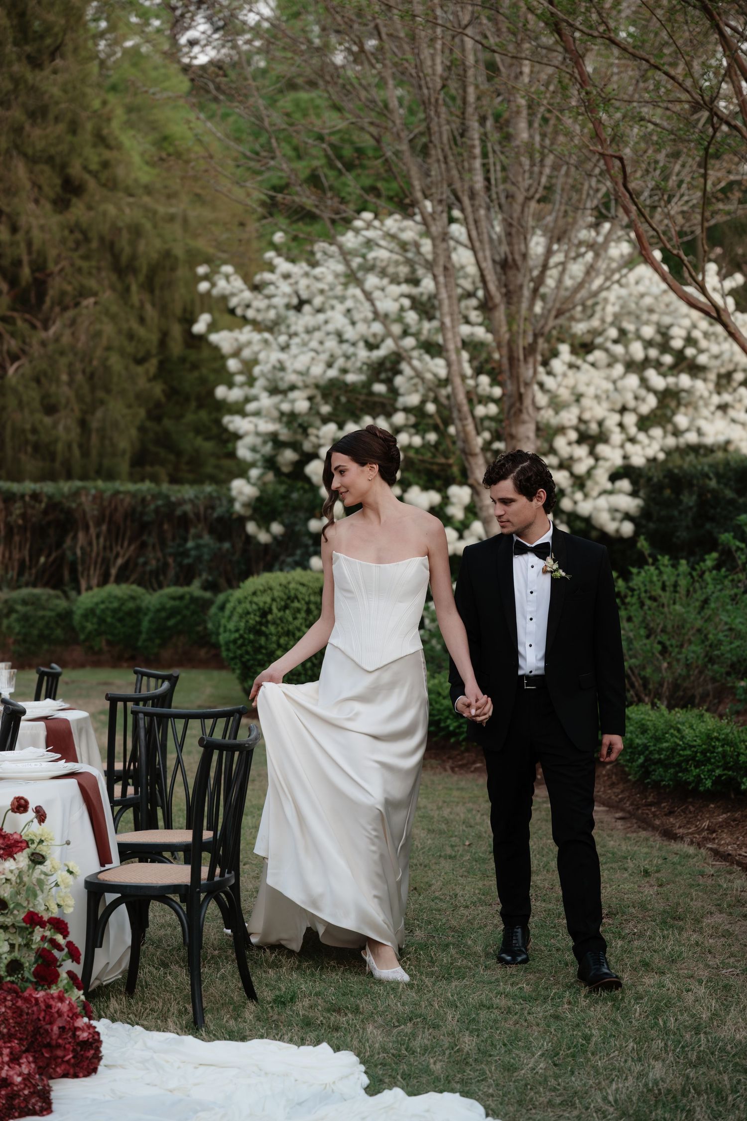 Bride in white gown and groom in black tuxedo walk hand in hand through lush garden at outdoor wedding reception.