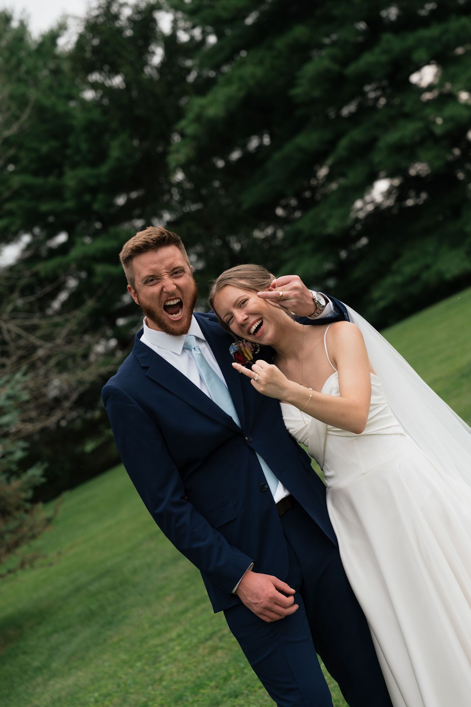 Joyful bride and groom laughing outdoors on green lawn, groom in navy suit, bride in white gown, lush trees behind.