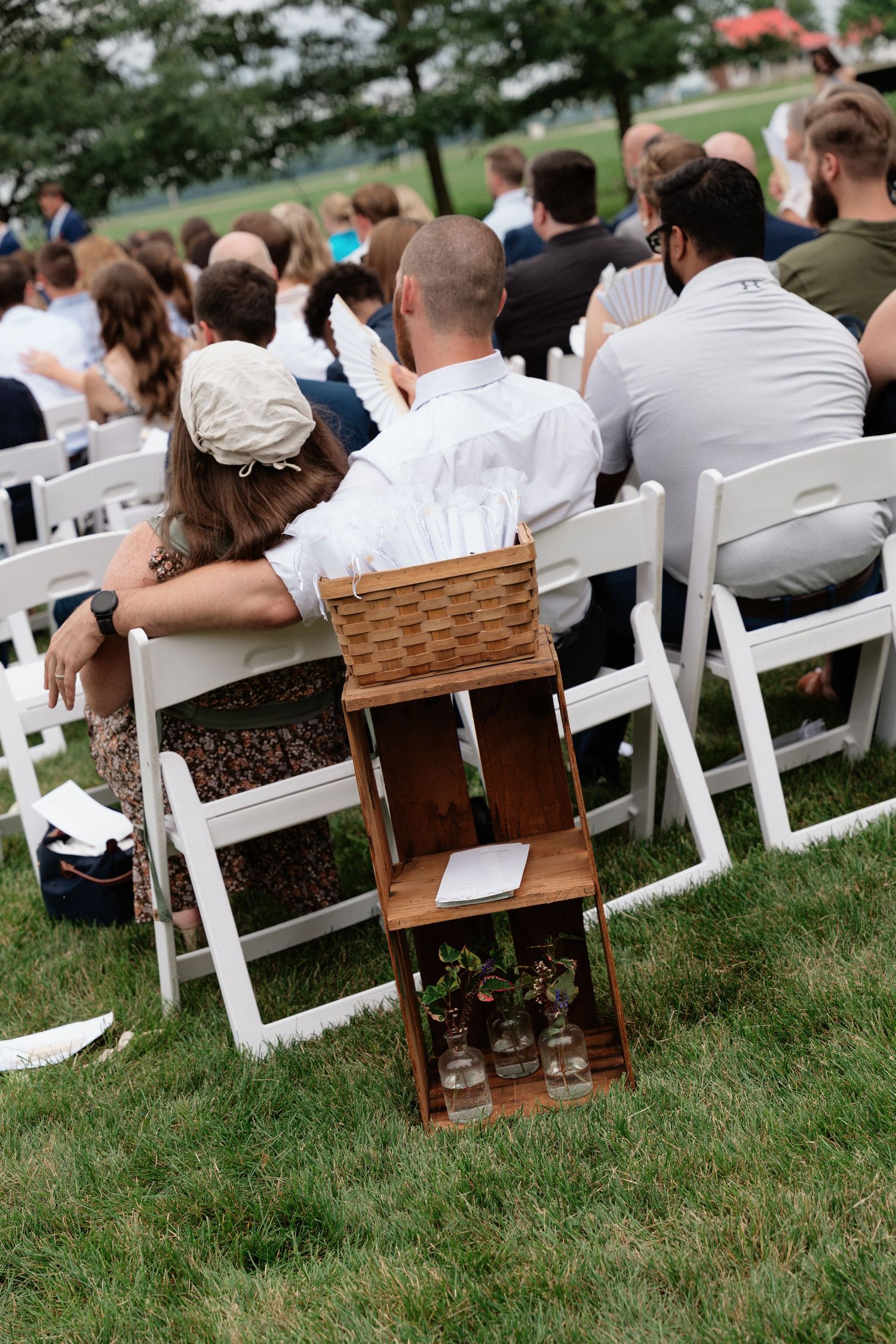 Guests seated in white chairs at an outdoor wedding ceremony on green grass with a rustic wooden crate decoration.