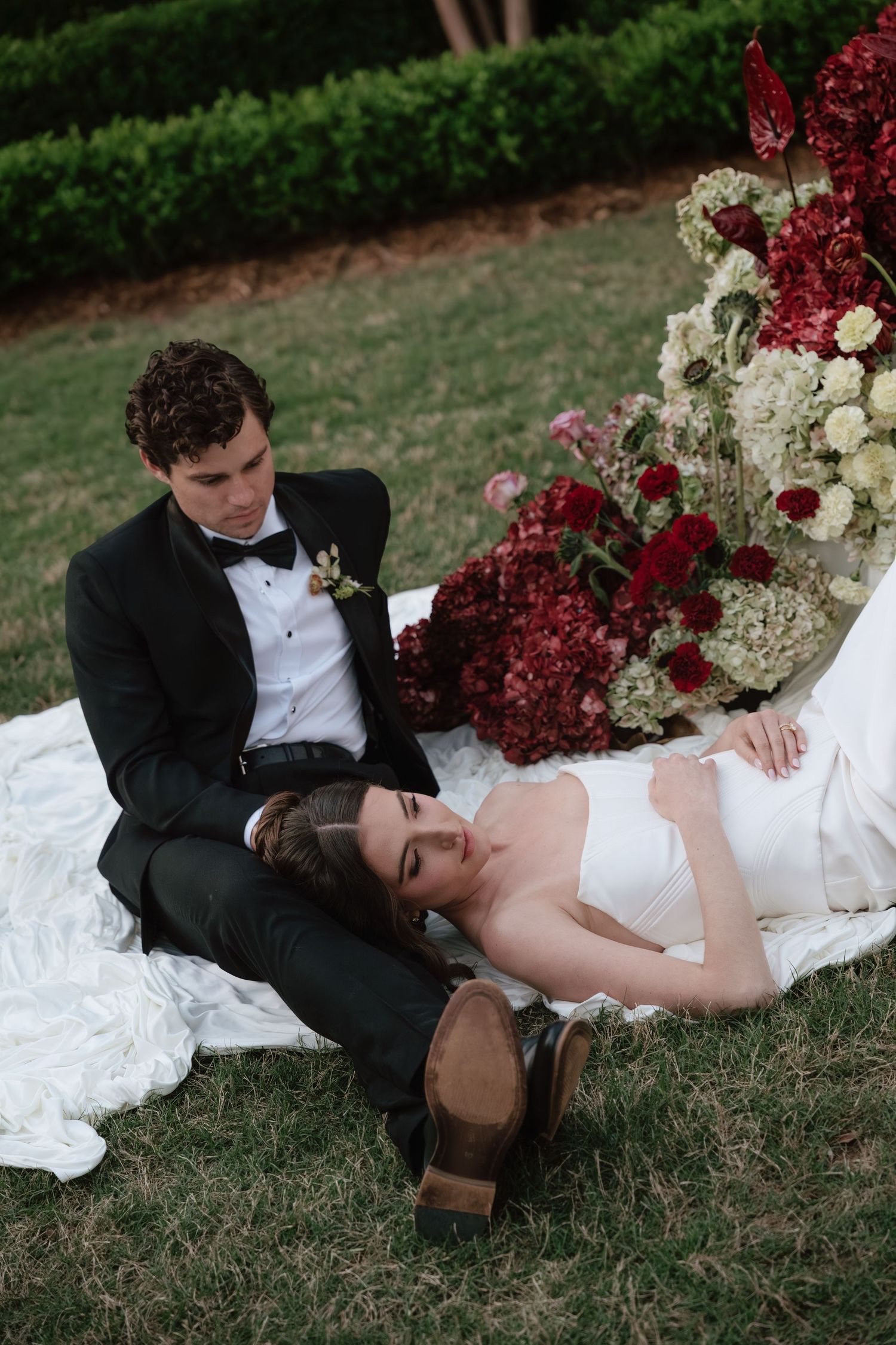 Bride in white gown rests on groom's lap outdoors near lush red and white floral arrangement on their wedding day.
