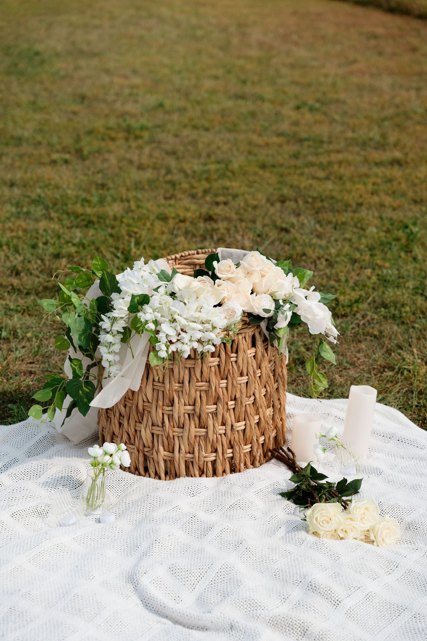 Rustic wicker basket filled with white roses and flowers on a white blanket outdoors on green grass.