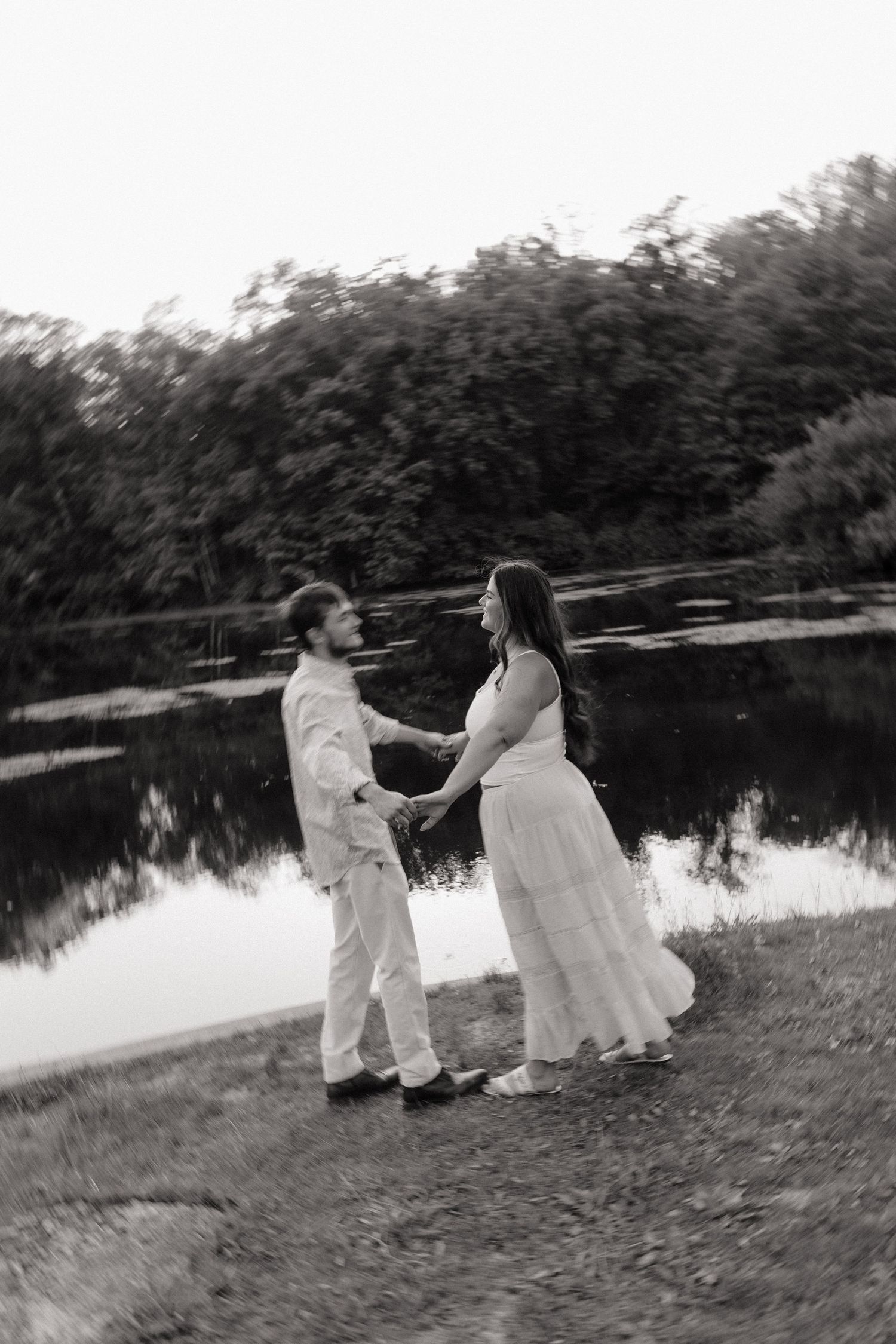 Black and white photo of a couple holding hands by a reflective pond, surrounded by lush trees.