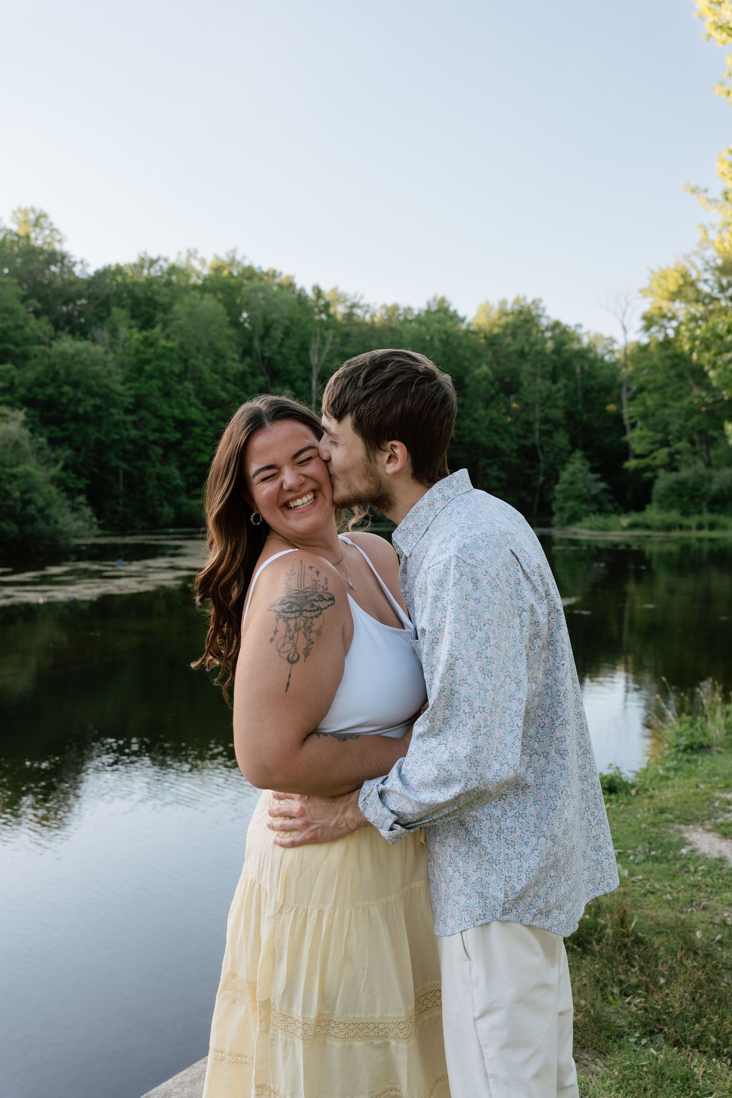 A couple embraces by a serene lake, woman smiling with tattoos, man kissing her cheek, surrounded by lush green trees.