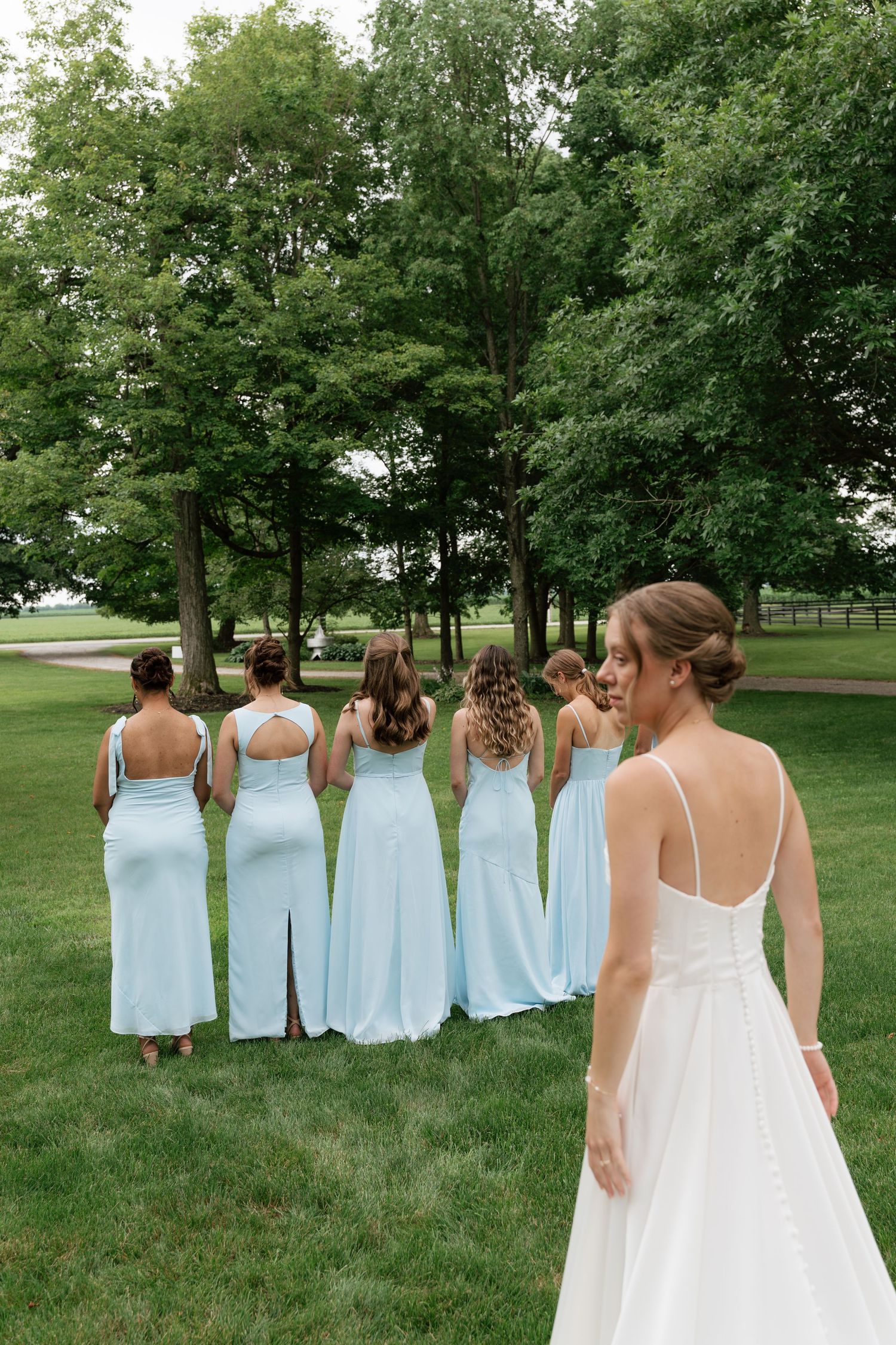 Bride in white gown stands with bridesmaids in light blue dresses on green lawn with trees in background.