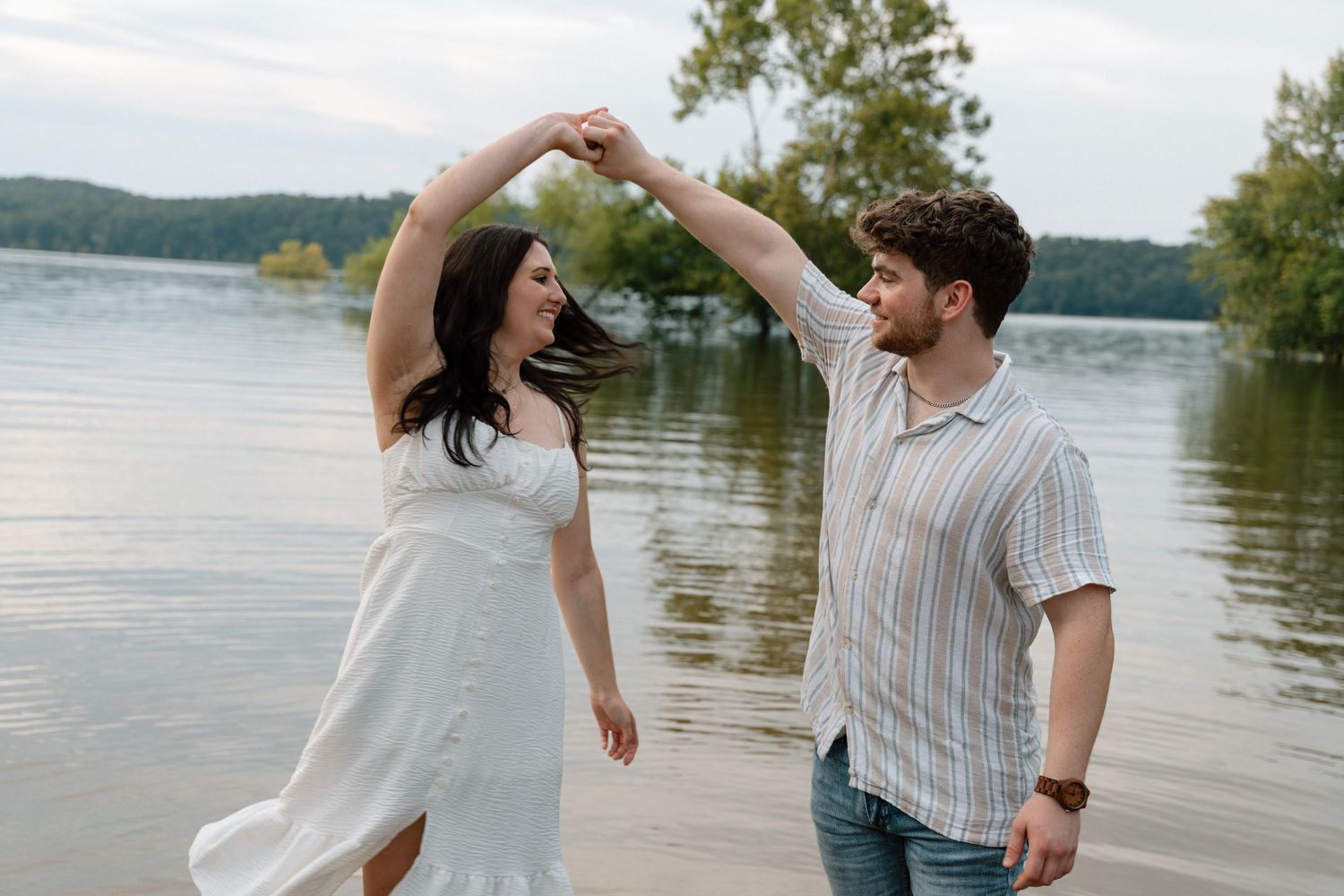 Couple dancing by a lakeside, woman in white dress being twirled by man in striped shirt, trees in background.