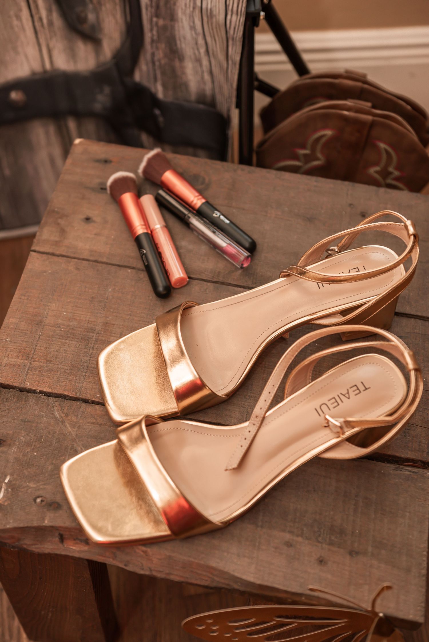 Rose gold strappy heeled sandals beside makeup brushes and lipstick on a rustic wooden table.