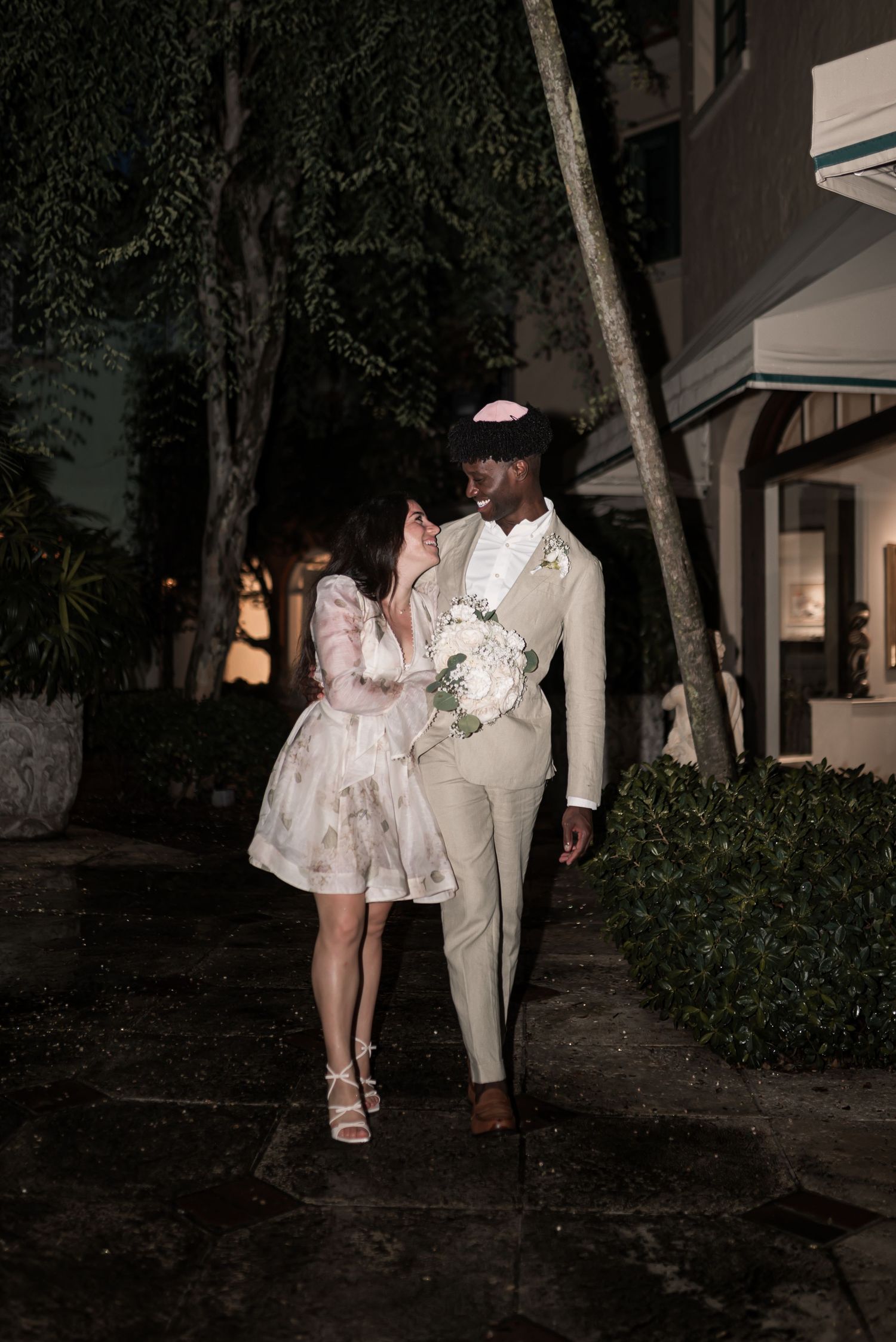 A couple walks along a dimly lit garden path at night wearing formal white attire.