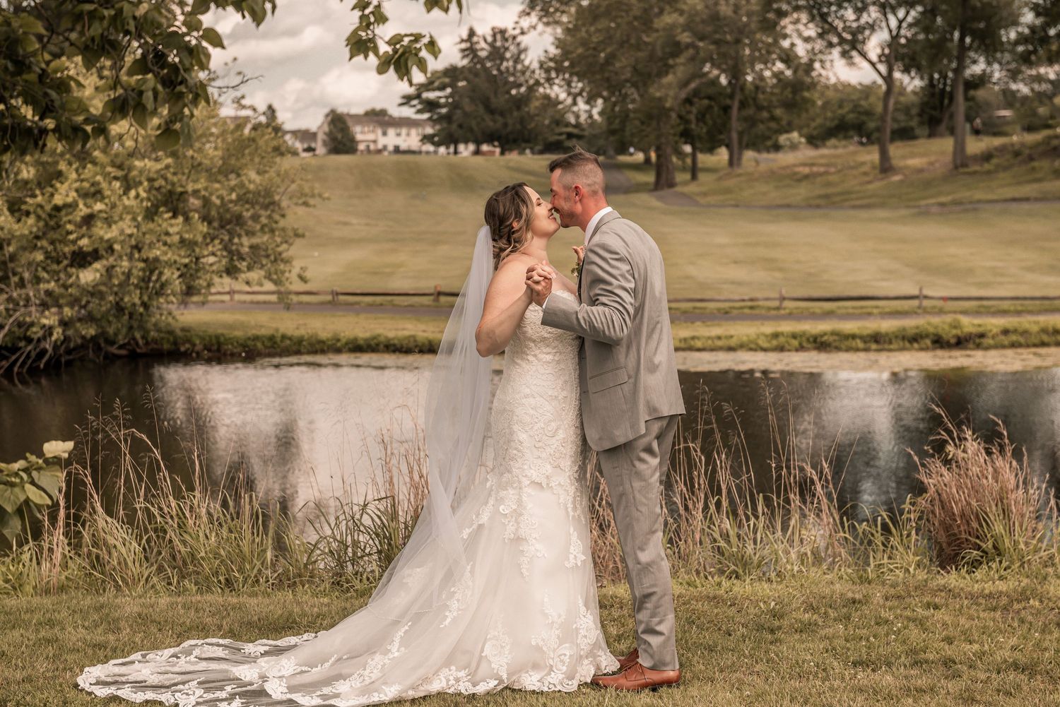 A romantic wedding couple shares an intimate moment beside a pond surrounded by autumn foliage and tall ornamental grasses.