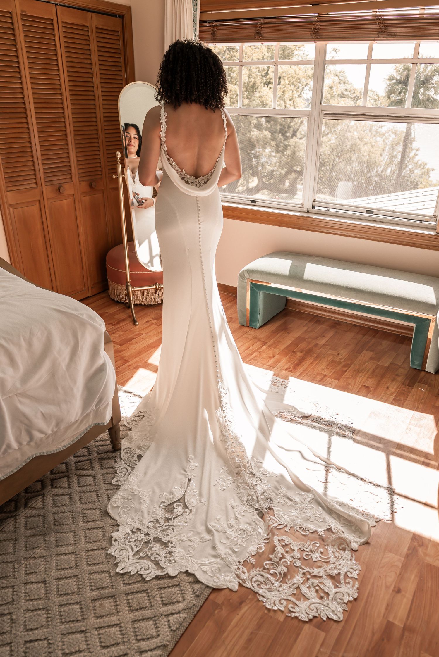 Bride in elegant backless lace wedding gown with train standing before mirror in sunlit room.