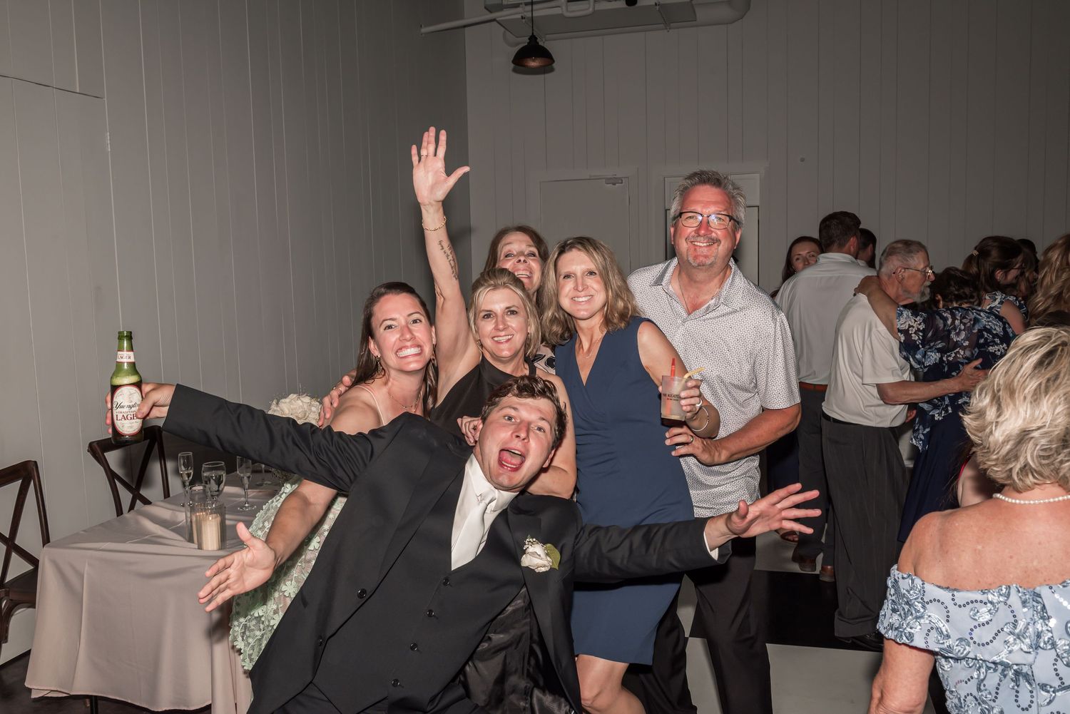 Group of friends celebrating at a wedding reception, posing and dancing joyfully in a bright indoor venue.