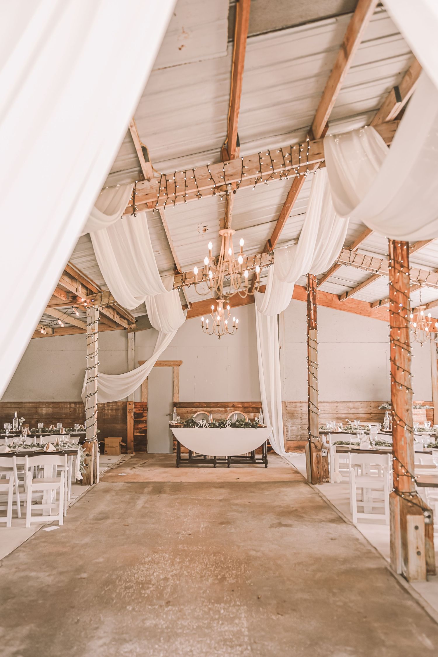 White draped fabric and string lights decorate a rustic barn venue interior with exposed wooden beams and concrete floors.