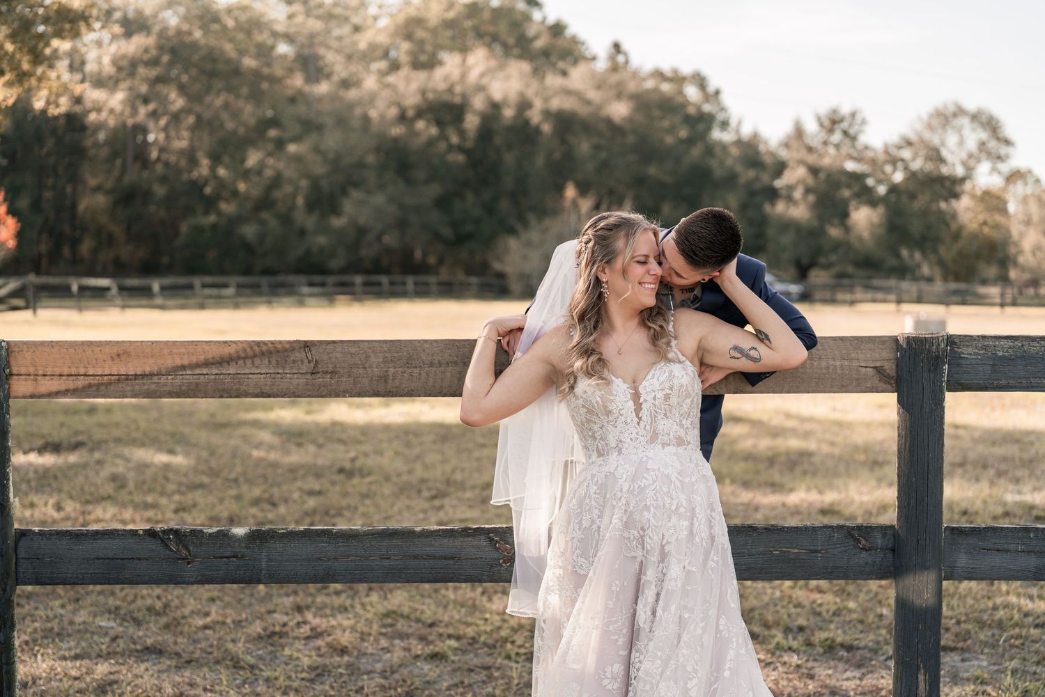 A couple in elegant white attire share an intimate moment in a rustic outdoor setting during golden hour.