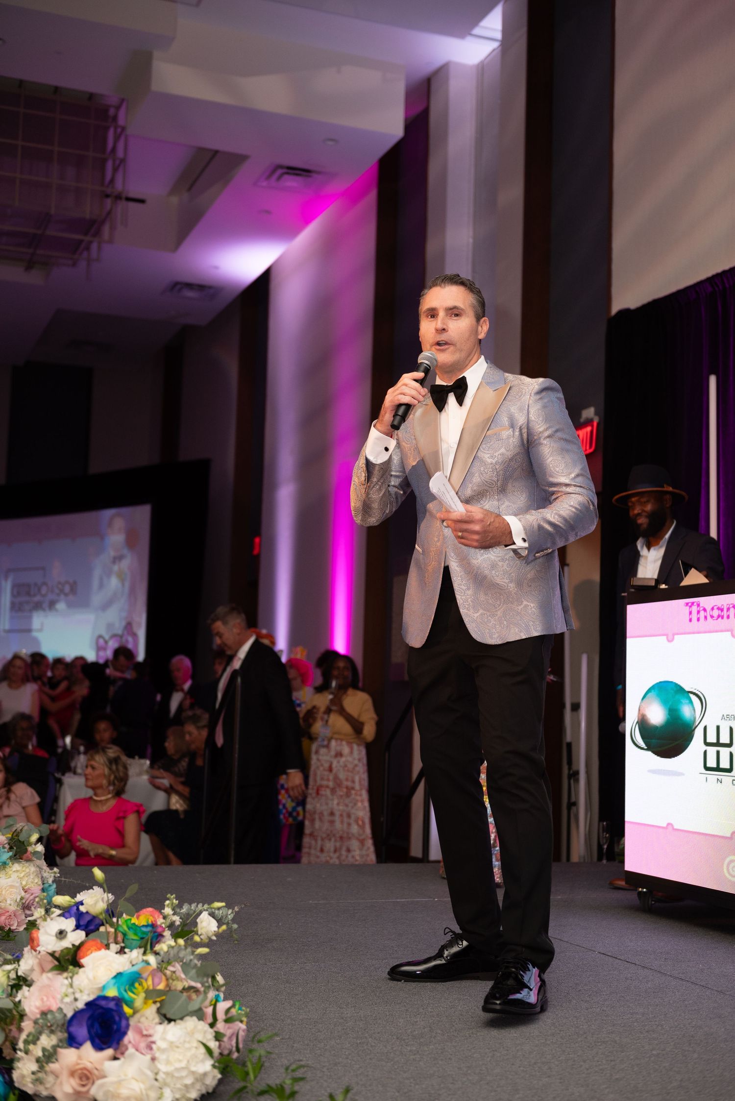 A performer in a silver suit jacket speaks into a microphone on stage at an elegant event with purple lighting and floral decor.