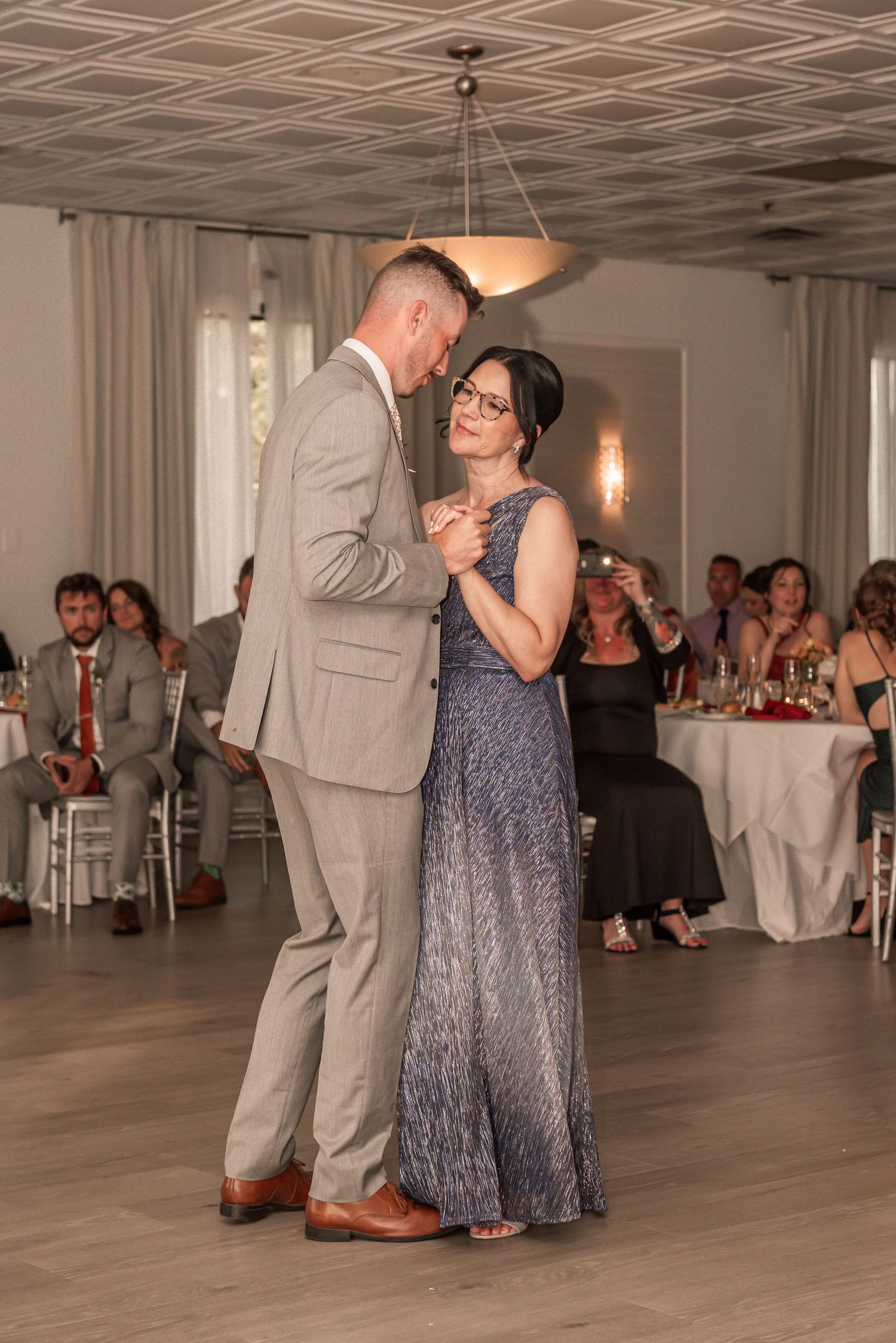 Dancers in formal attire, including a grey suit and blue dress, perform elegant dance moves on a wooden floor.