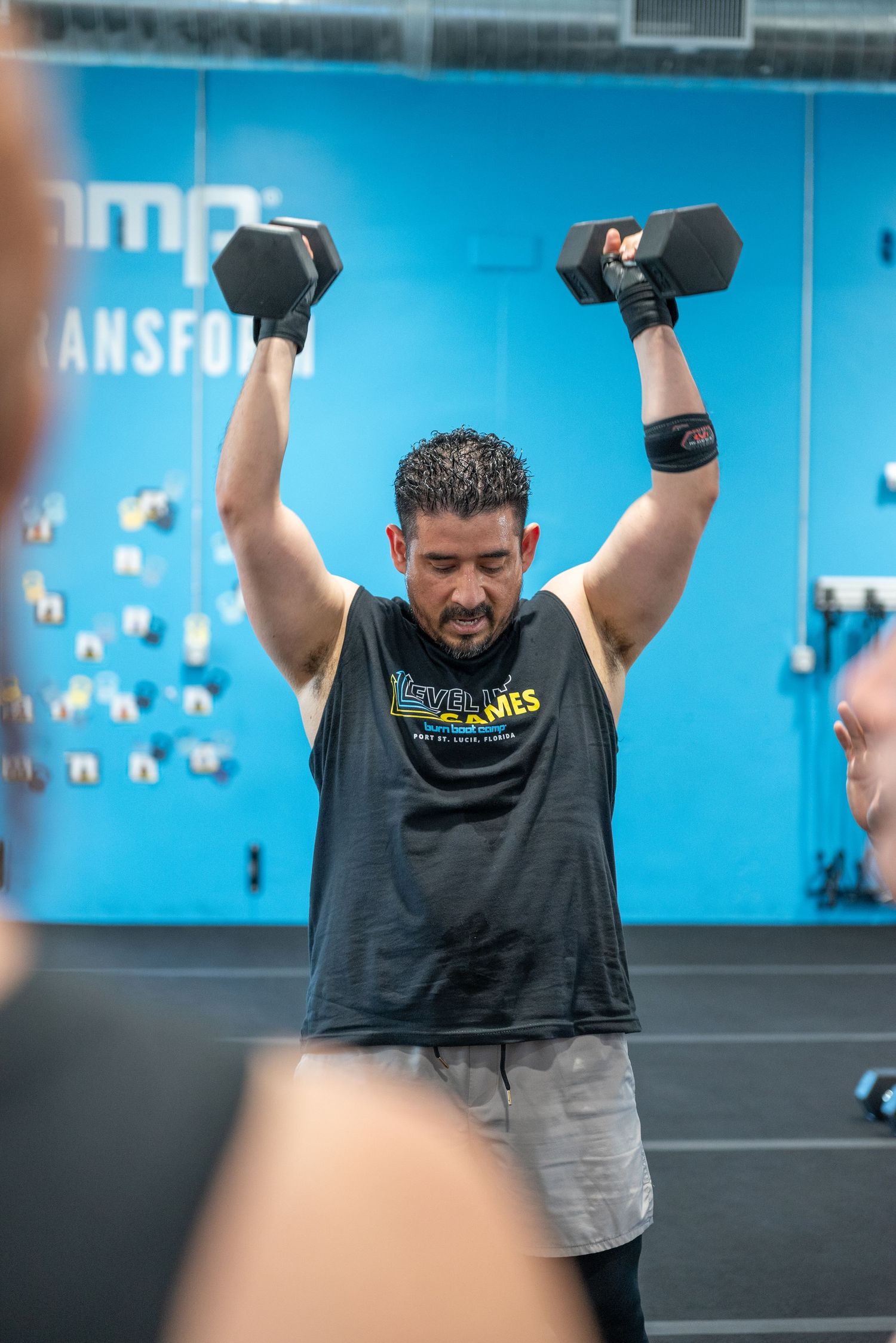 Man lifting dumbbells overhead in a gym wearing a black tank top during a fitness workout session.