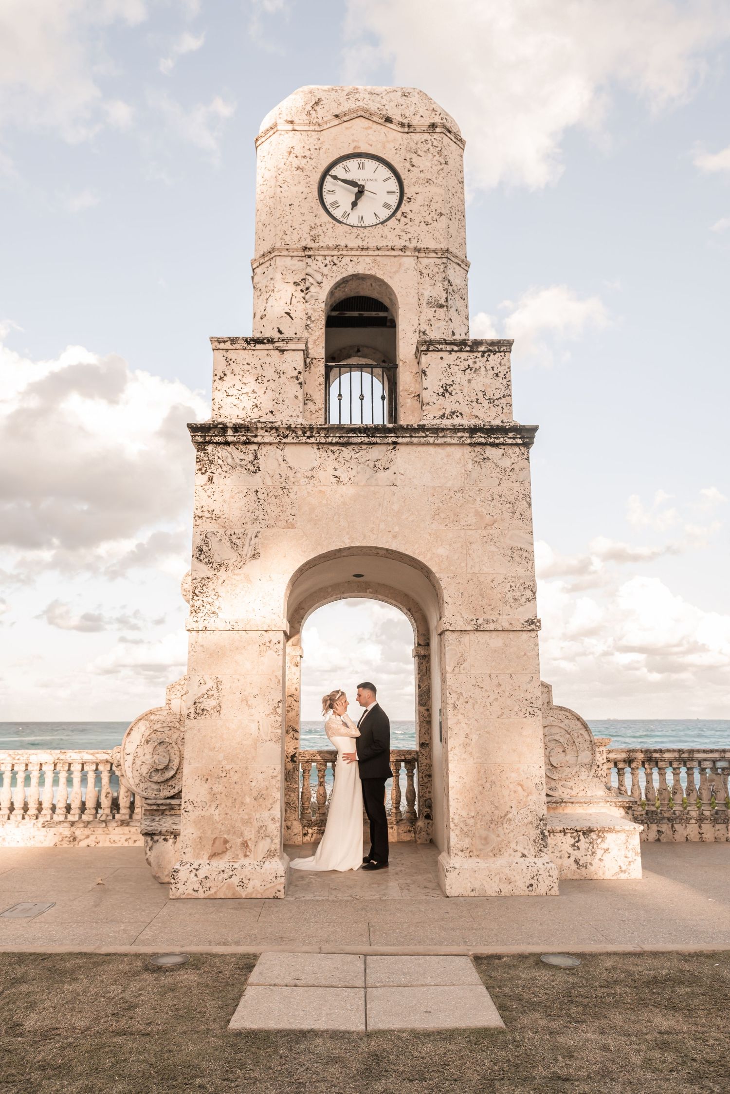 Bride and groom share a romantic moment under a historic clock tower arch with ocean views in Palm Beach.