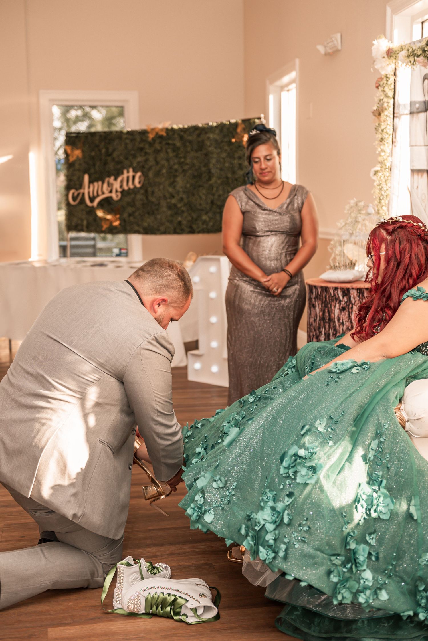 Man kneeling to place shoes on a young woman in a teal quinceañera gown during a ceremony with name backdrop.