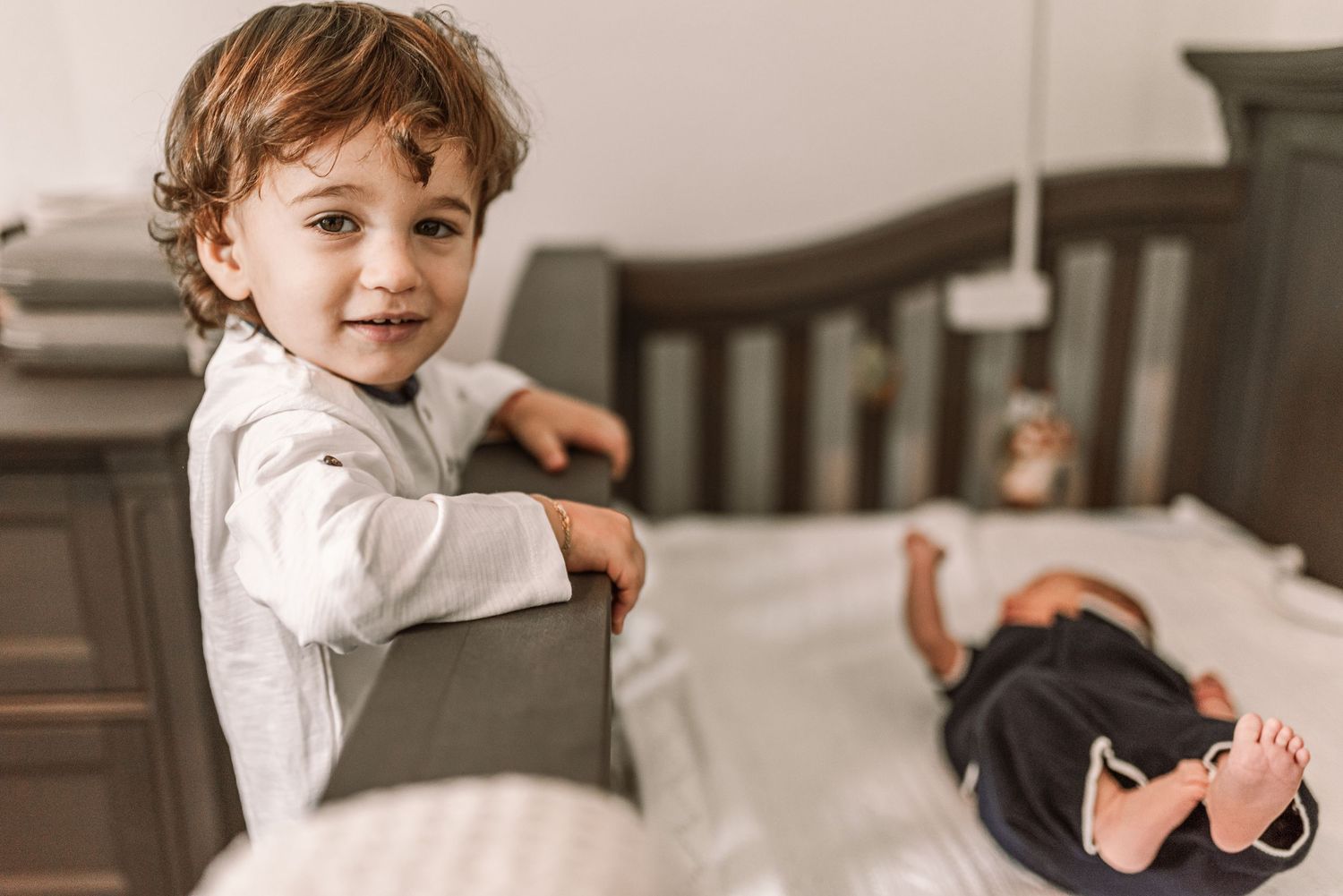 Smiling toddler stands by a crib next to a sleeping newborn baby in a cozy nursery bedroom.