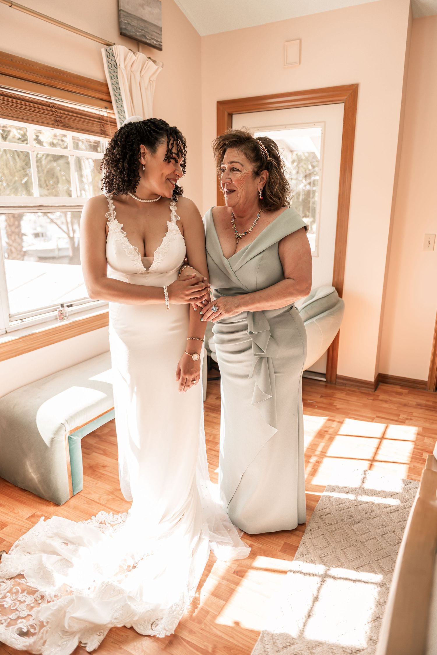 Bride in white lace gown smiles with older woman in sage dress while getting ready indoors.