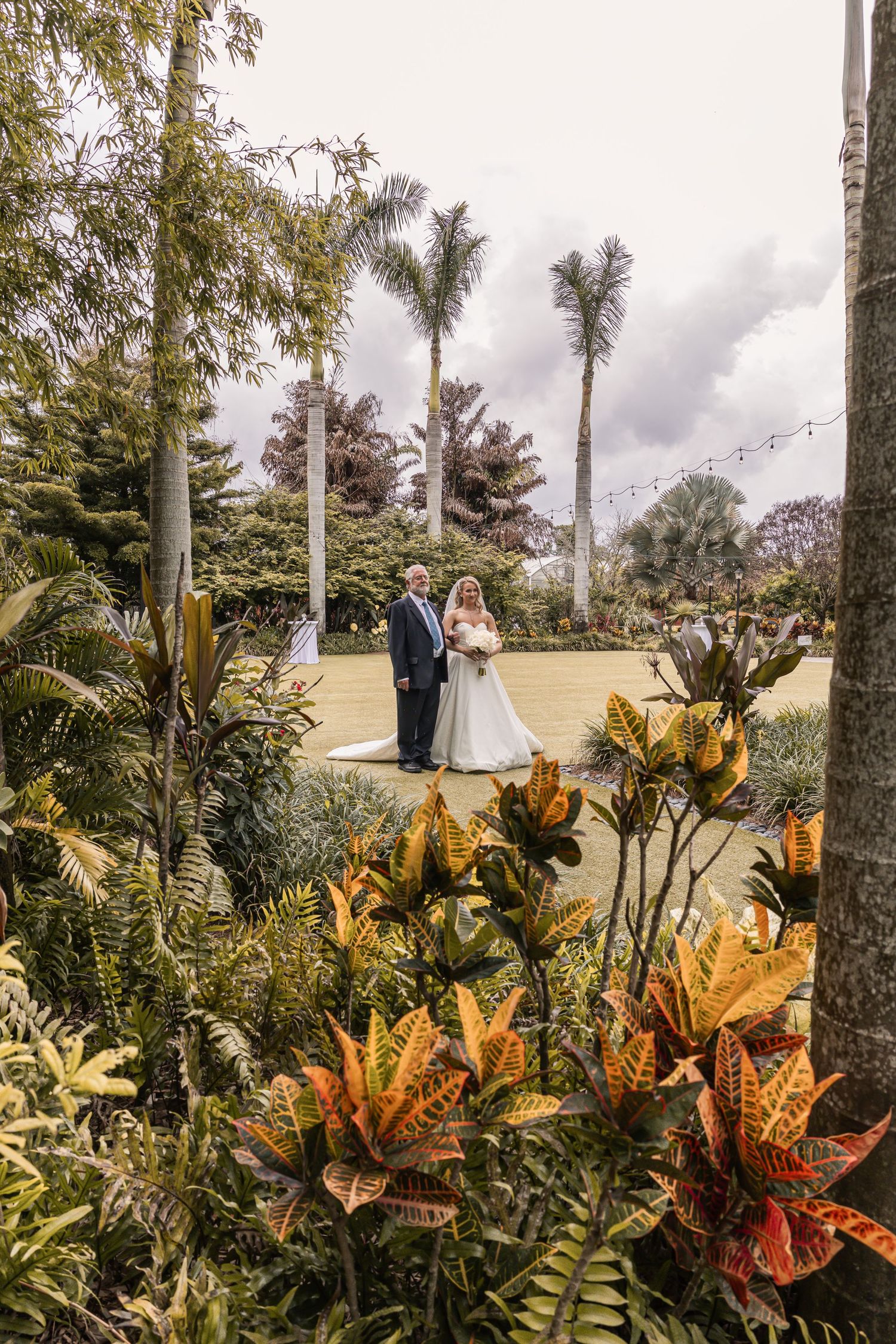 Bride and groom posing in a lush tropical garden with palm trees and colorful croton plants on their wedding day.