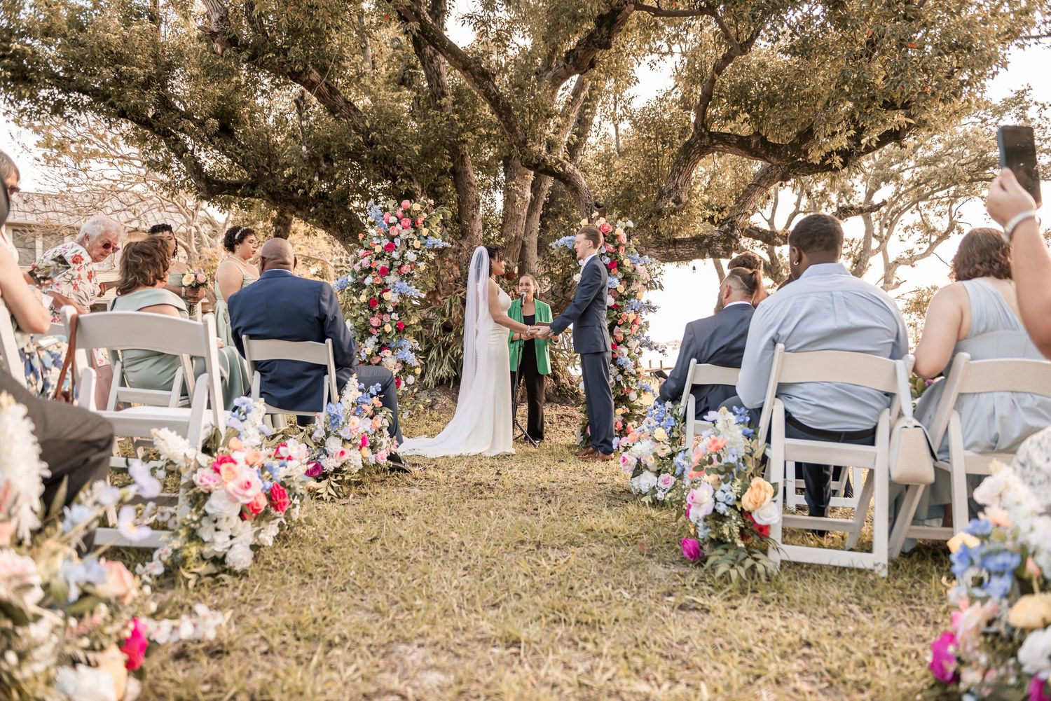 Outdoor wedding ceremony under a large oak tree with colorful floral arch and aisle arrangements, guests seated in white chairs.