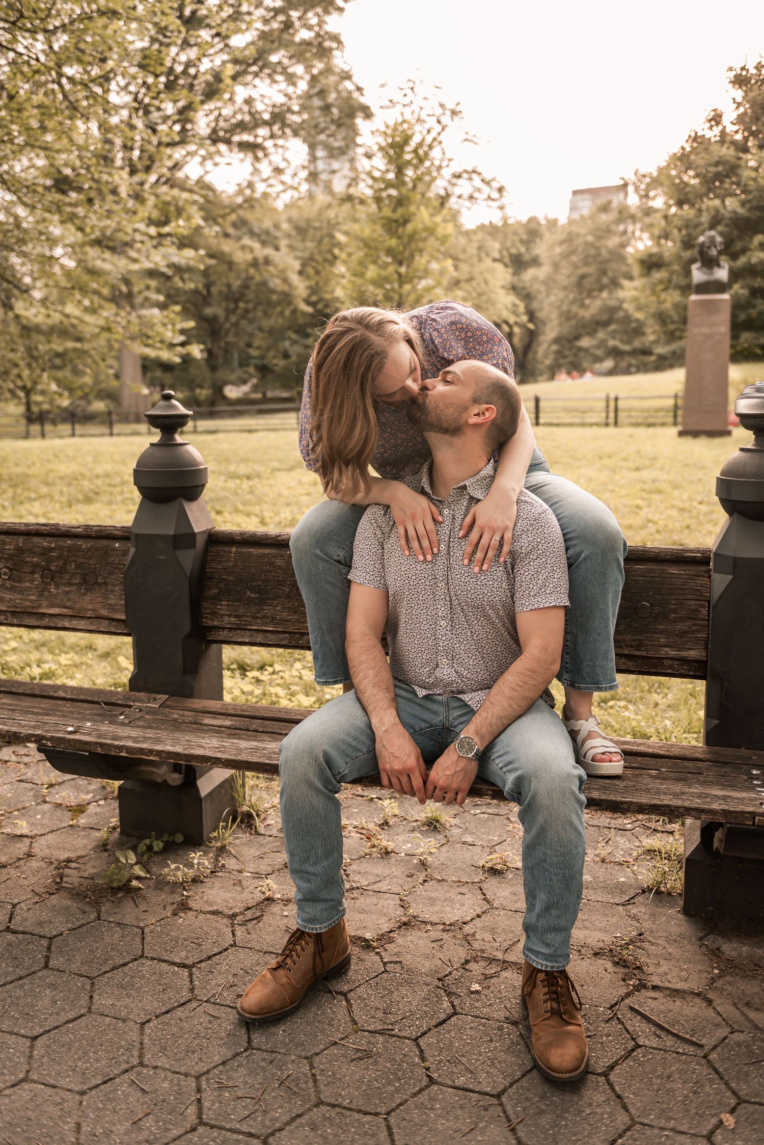 A romantic couple shares an intimate moment on a park bench surrounded by autumn trees and stone pavers.