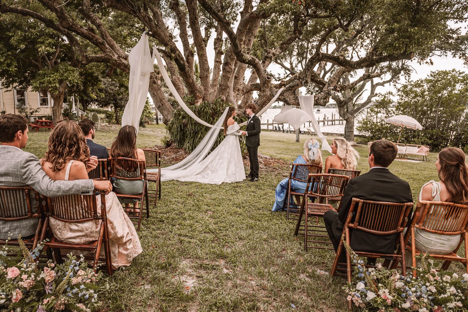 Rustic outdoor wedding ceremony setup with wooden chairs arranged in rows under large trees at sunset.