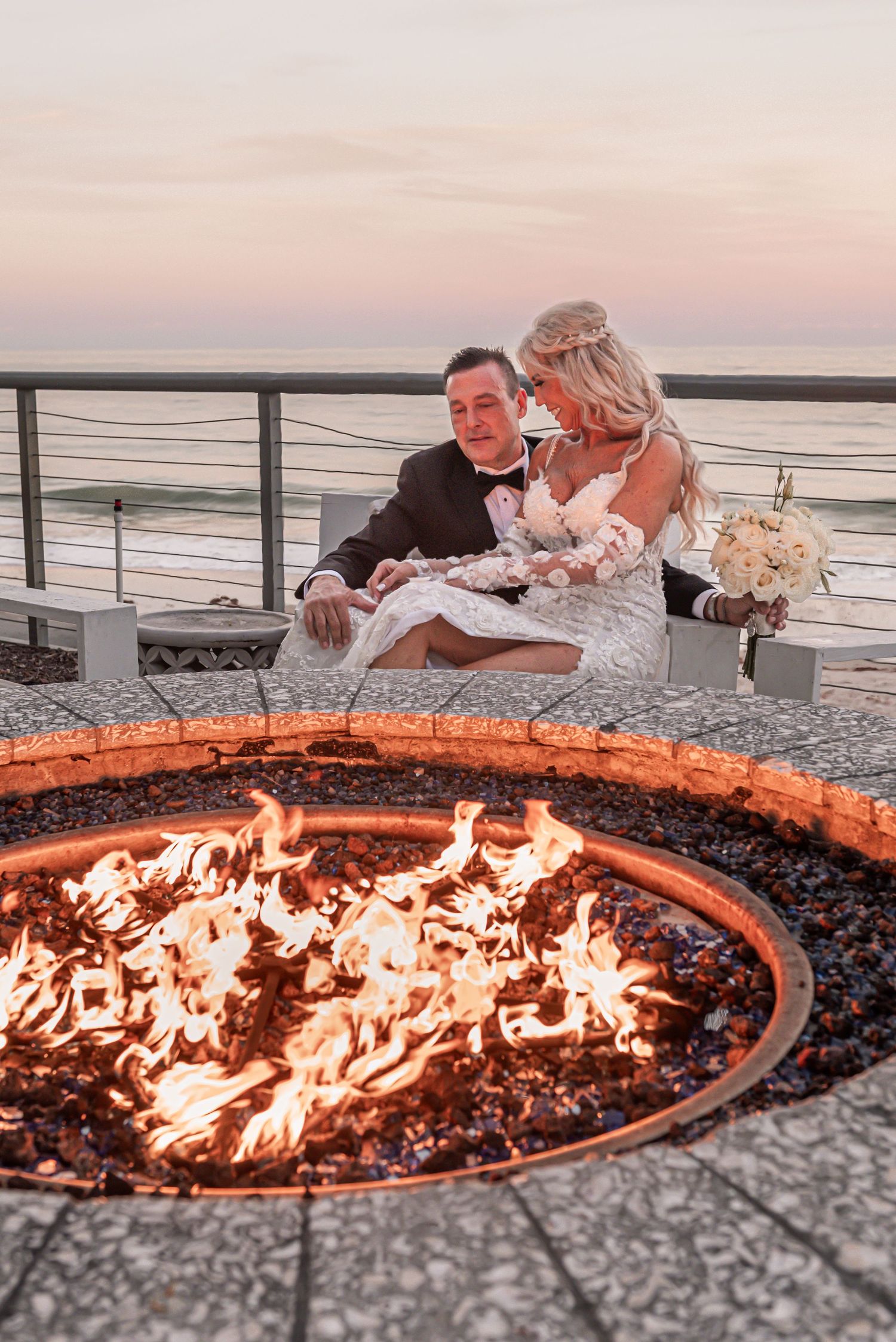 Couple relaxing by a glowing fire pit at sunset overlooking the ocean from a beachside patio.
