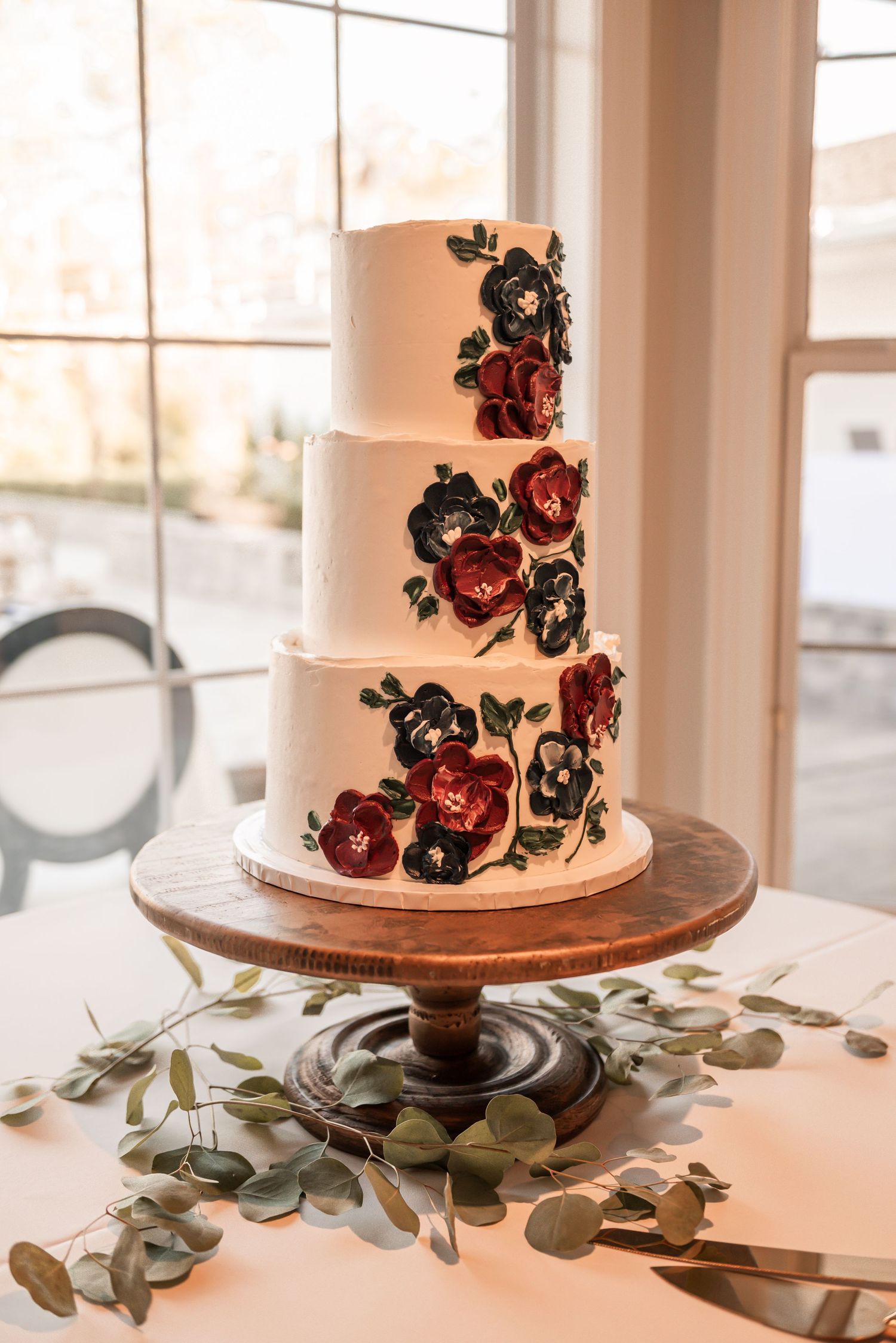 Elegant white wedding cake adorned with red and black sugar flowers on wooden cake stand with eucalyptus leaves.